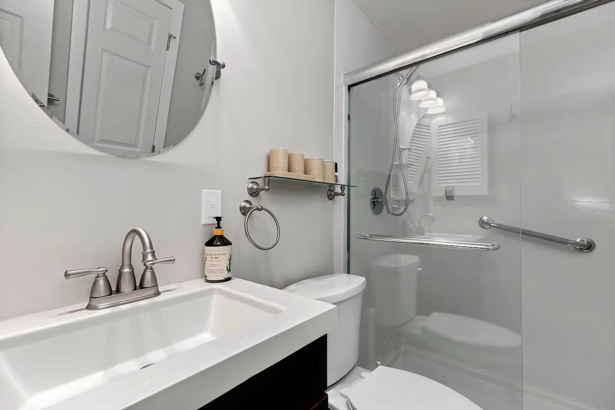 A small bathroom with a white sink, a wall-mounted mirror, a soap dispenser, a toilet, and a glass shower enclosure with a showerhead. There is a shelf with candles above the toilet and a hand towel ring next to the mirror.