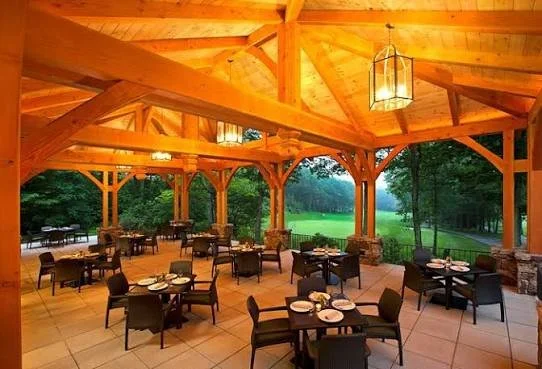 Upscale outdoor patio dining area with wooden beams, hanging lanterns, tables set with plates and utensils, overlooking a lush green landscape.