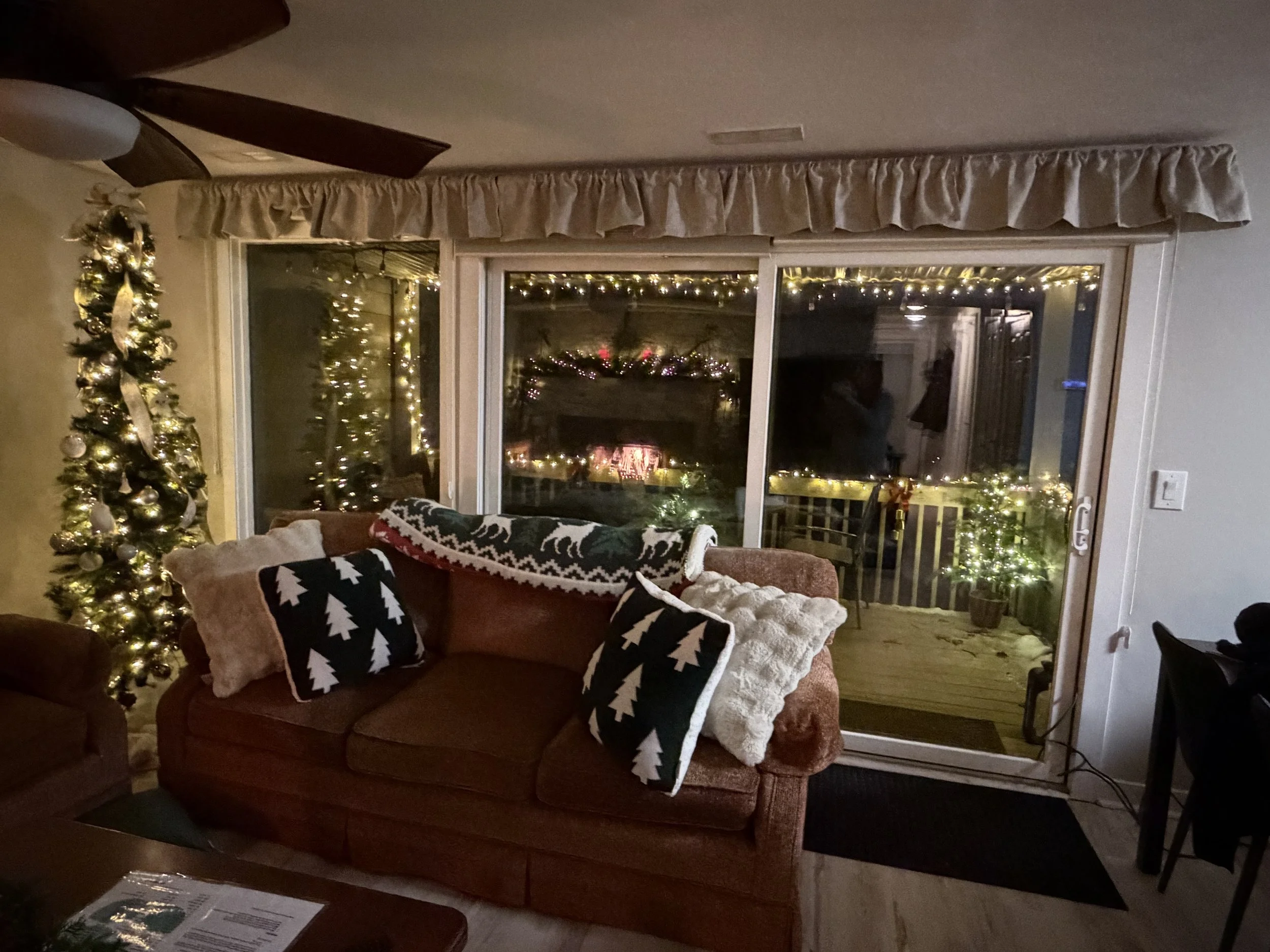 Living room decorated for Christmas with a lit Christmas tree in the corner, a couch with holiday-themed pillows, a blanket with reindeer and snowflakes, and a sliding glass door leading to a deck outside decorated with fairy lights.