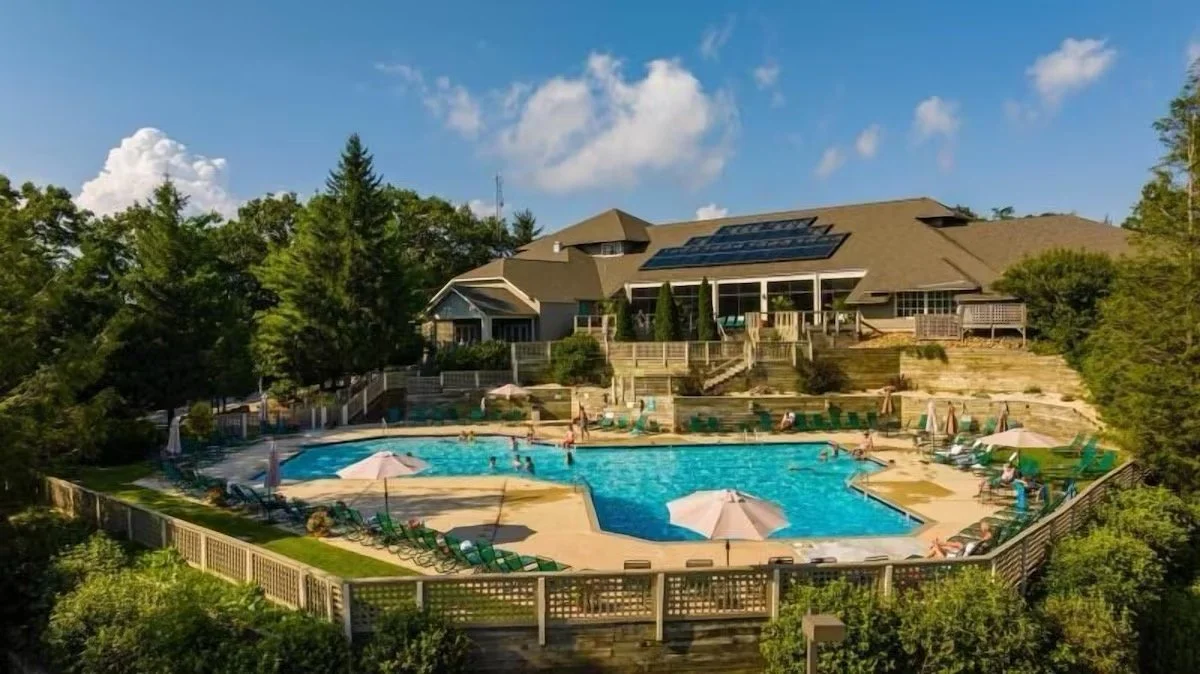 A large outdoor swimming pool surrounded by lounge chairs, umbrellas, and trees, with a large house or clubhouse in the background under a partly cloudy sky.