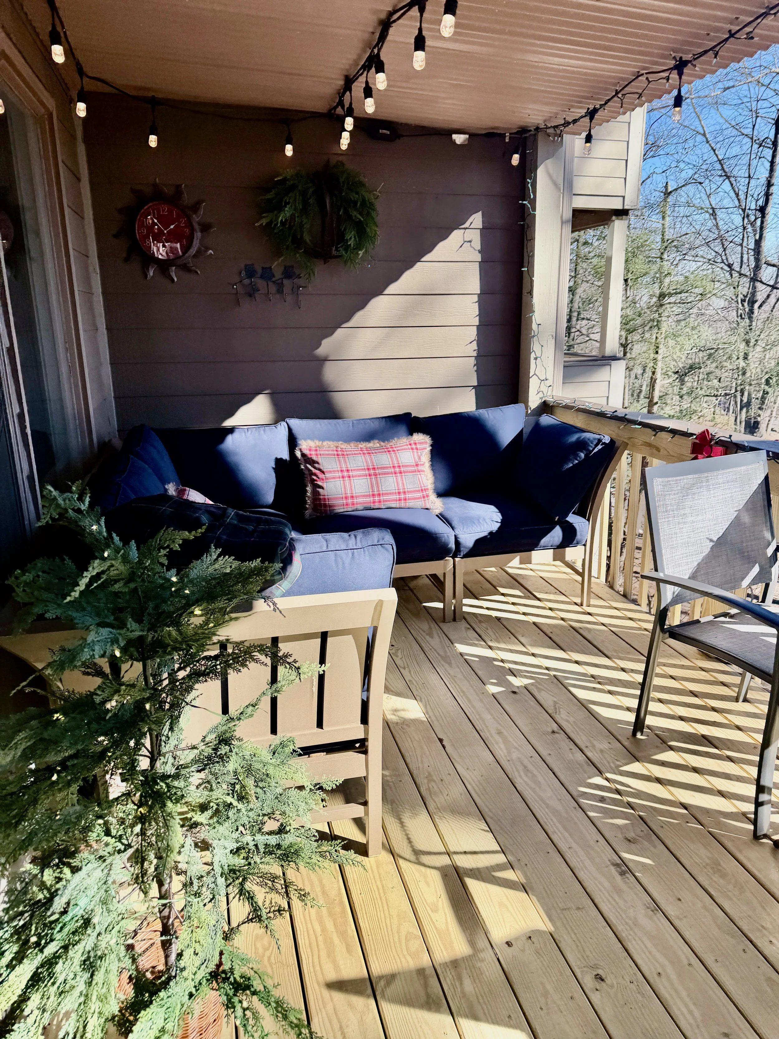 A cozy outdoor porch decorated for the holidays with string lights, a small Christmas tree, and a flower wreath, featuring cushioned seating.