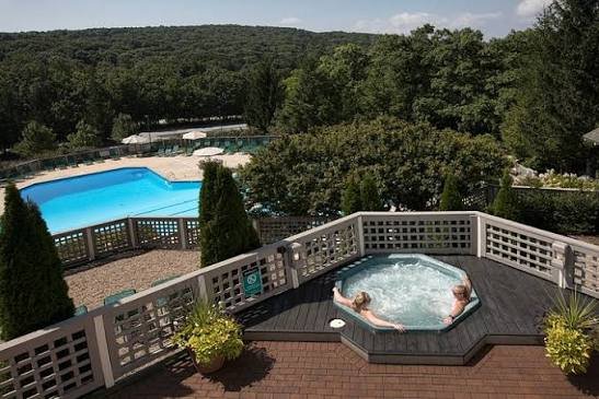 People in a hot tub on a wooden deck overlooking a swimming pool and lush green trees in the background.