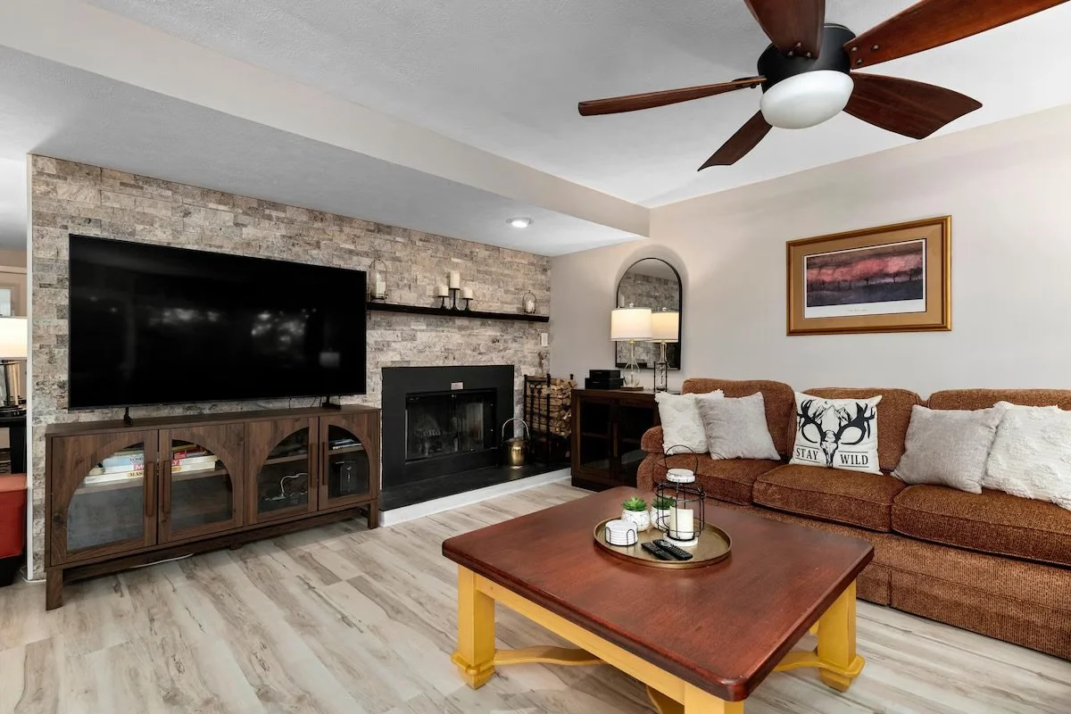 Living room with a brown couch, decorative pillows, a wooden coffee table, a brick fireplace, a mounted flat-screen TV, a ceiling fan, and a side table with a lamp.