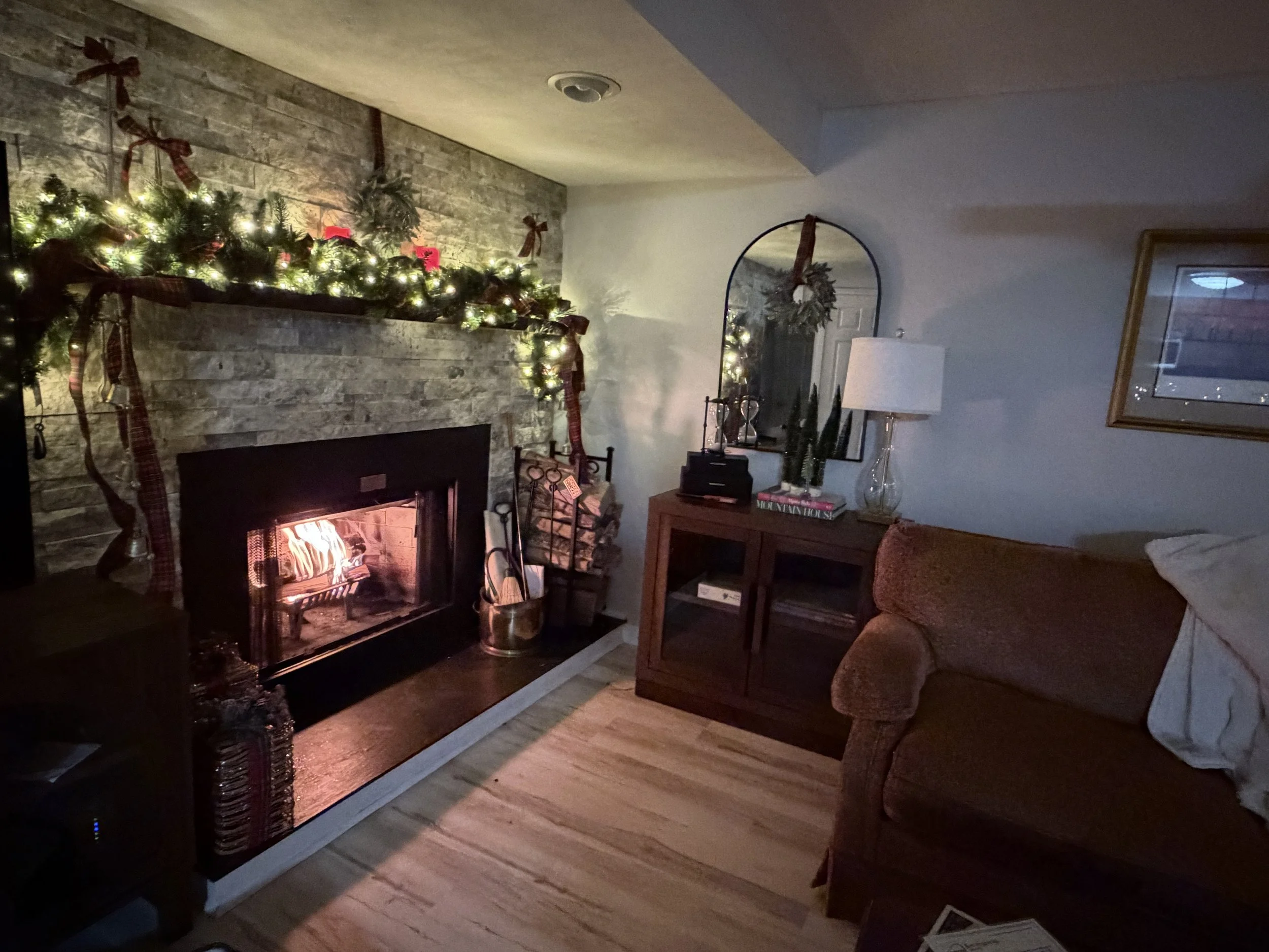 Cozy living room with lit fireplace decorated for Christmas. Mantel has green garland with lights, bows, and ornaments. Furniture includes a sofa, a wooden cabinet with books, a lamp, and a mirror with holiday wreath.