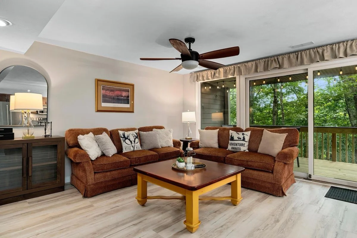 Living room with two brown sofas, decorative pillows, wooden coffee table, sliding glass door with greenery outside, ceiling fan, and a side lamp