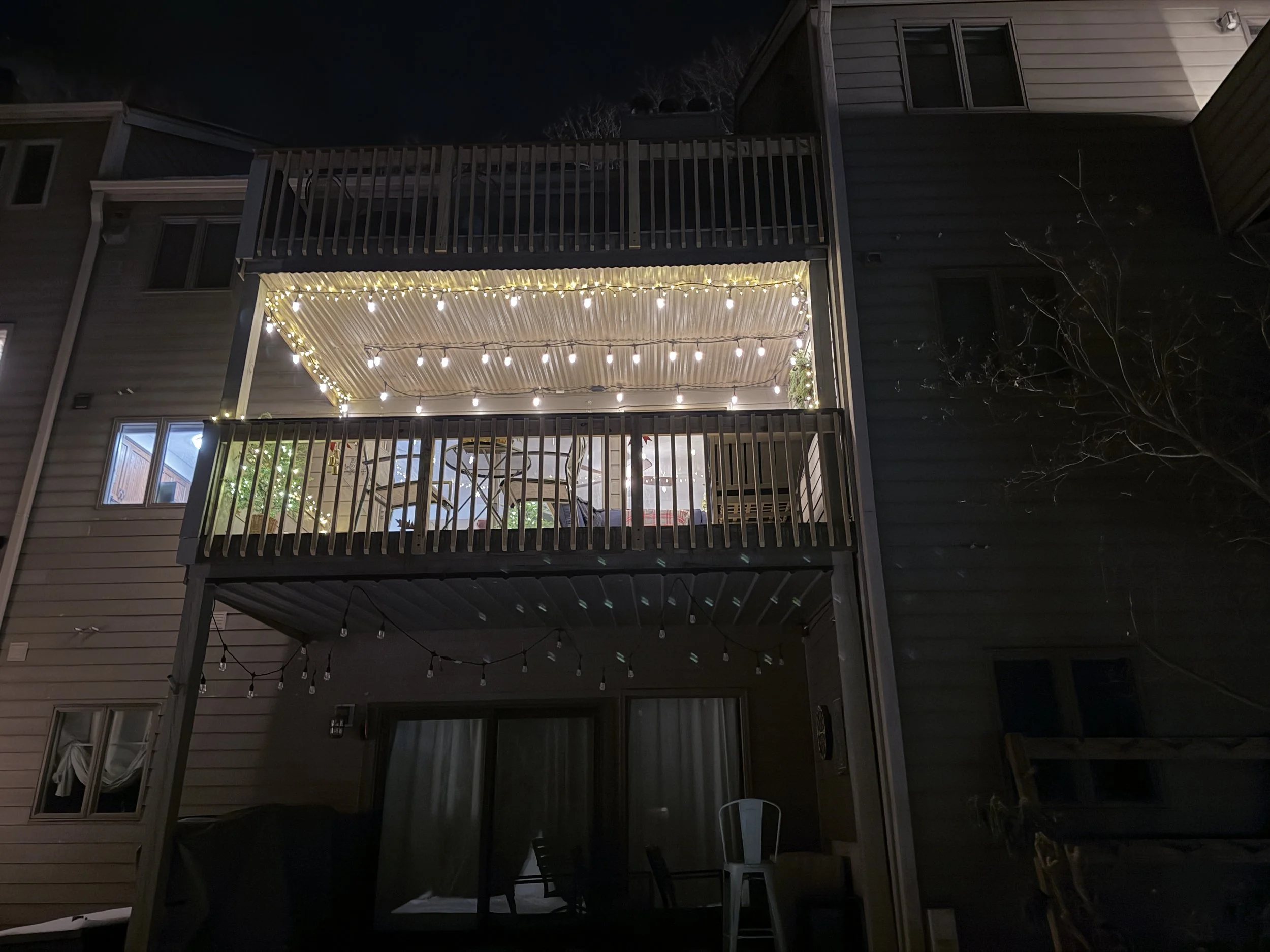 Nighttime view of a three-story house with illuminated balcony decorated with string lights, showing a dining area and outdoor furniture.