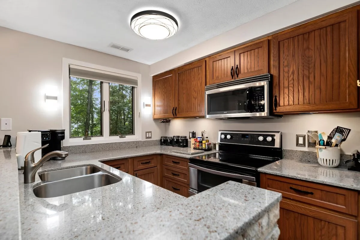 Kitchen with granite countertops, wooden cabinets, stainless steel microwave and oven, window with forest view, ceiling light fixture.