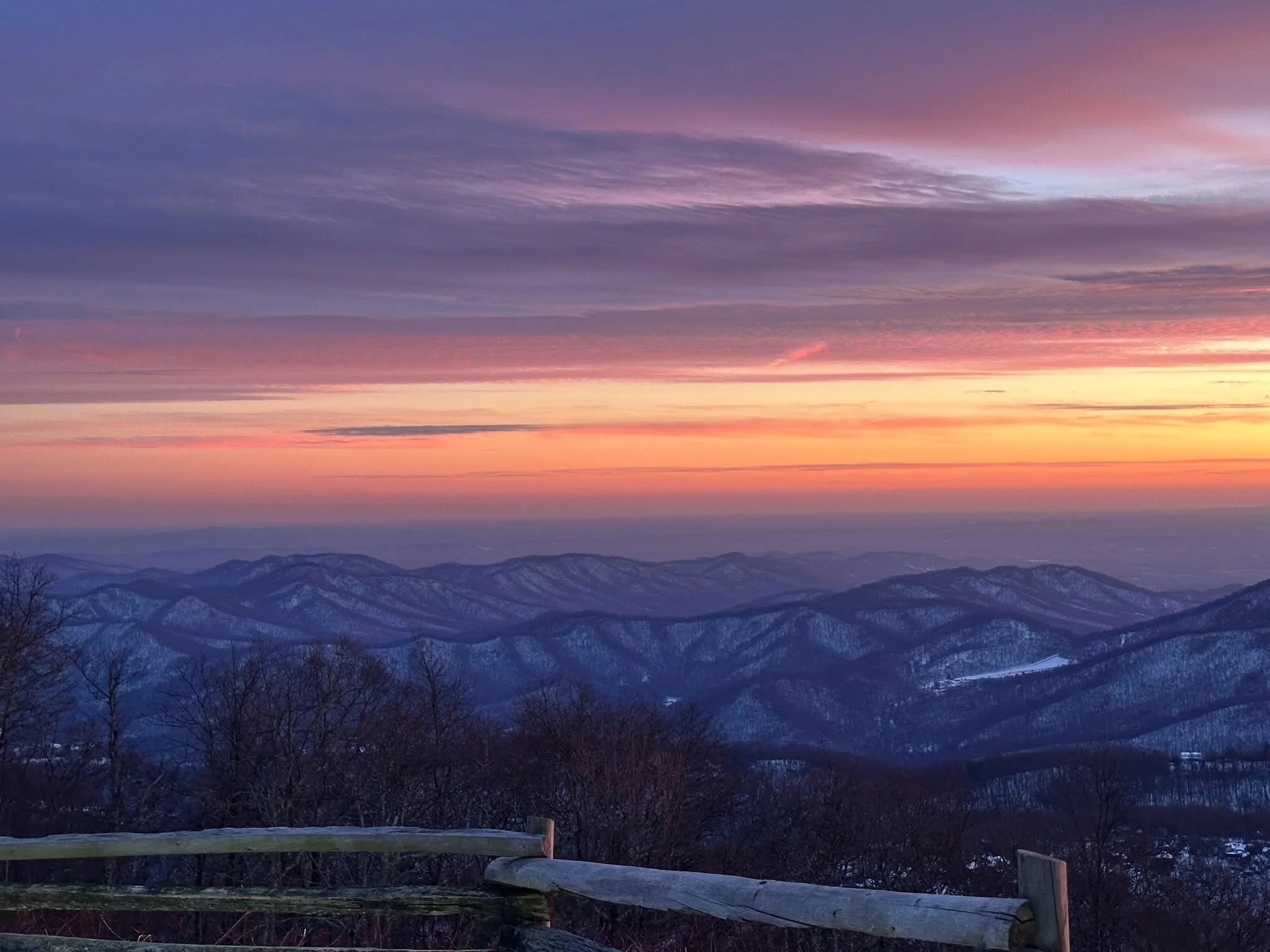 Sunset over snow-covered mountains with colorful sky and a wooden fence in the foreground.