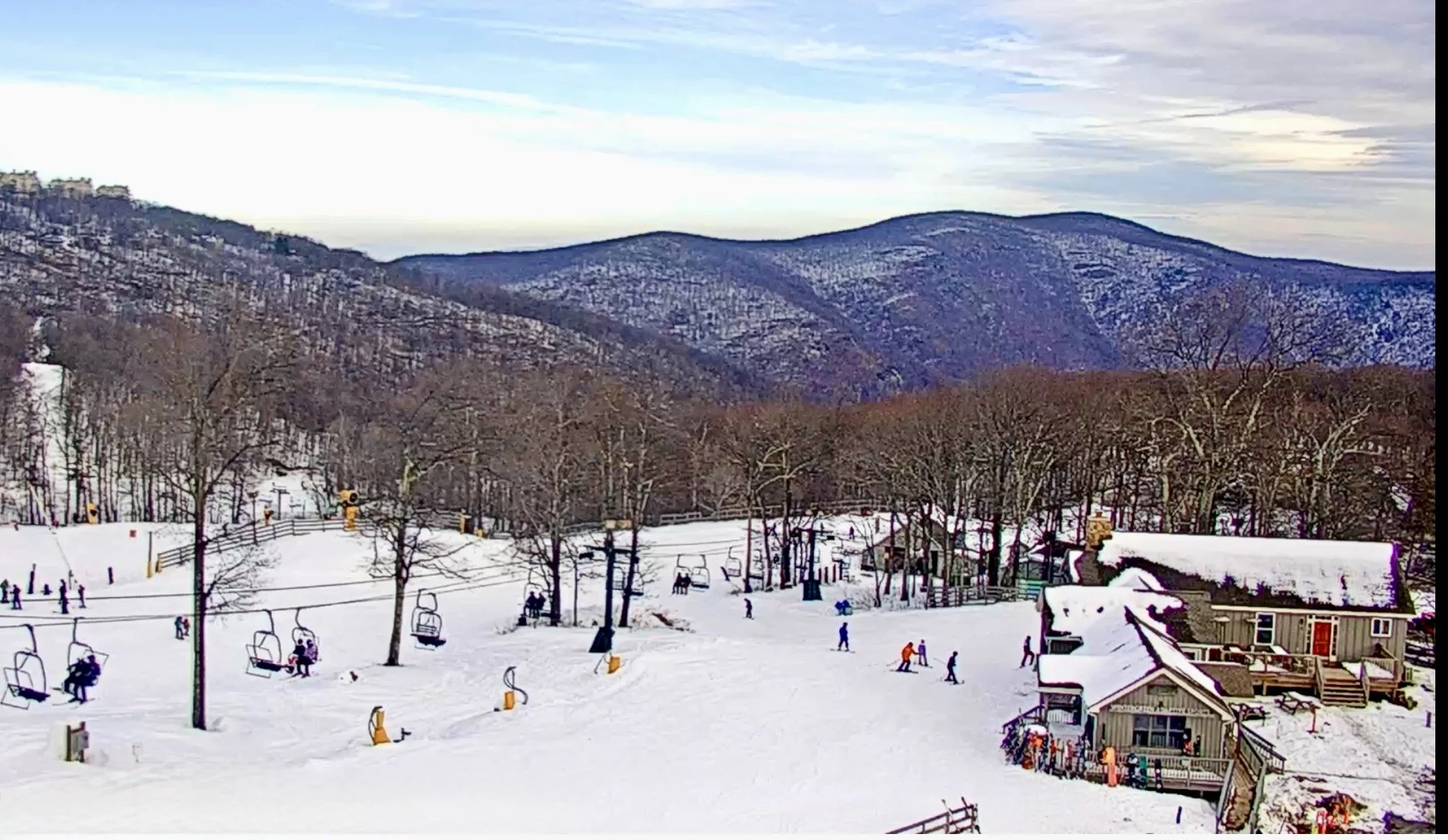 Ski resort with ski lifts, skiers, and snow-covered buildings surrounded by leafless trees and mountains in the background under a cloudy sky.