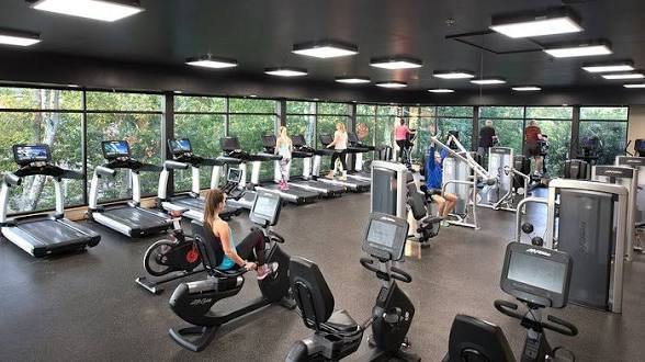 People working out on treadmills and exercise bikes in a gym with large windows and natural light.