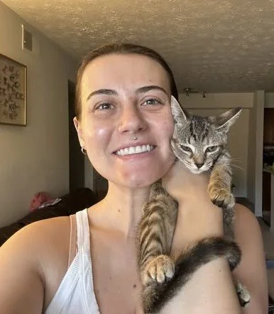 Woman smiling while holding a gray tabby cat indoors.