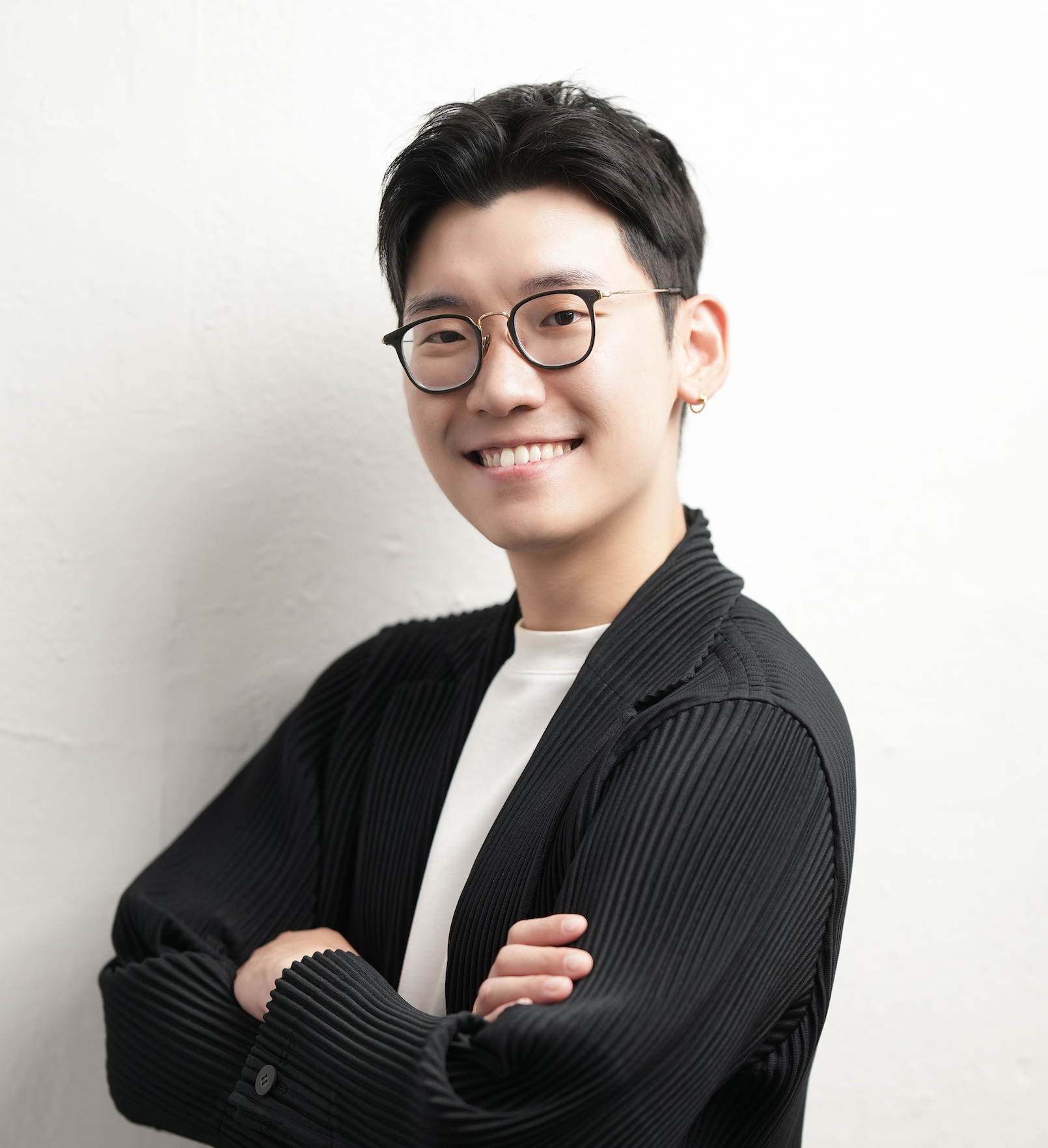 A young man wearing glasses, a black textured blazer, and a white t-shirt, smiling with arms crossed against a white wall.