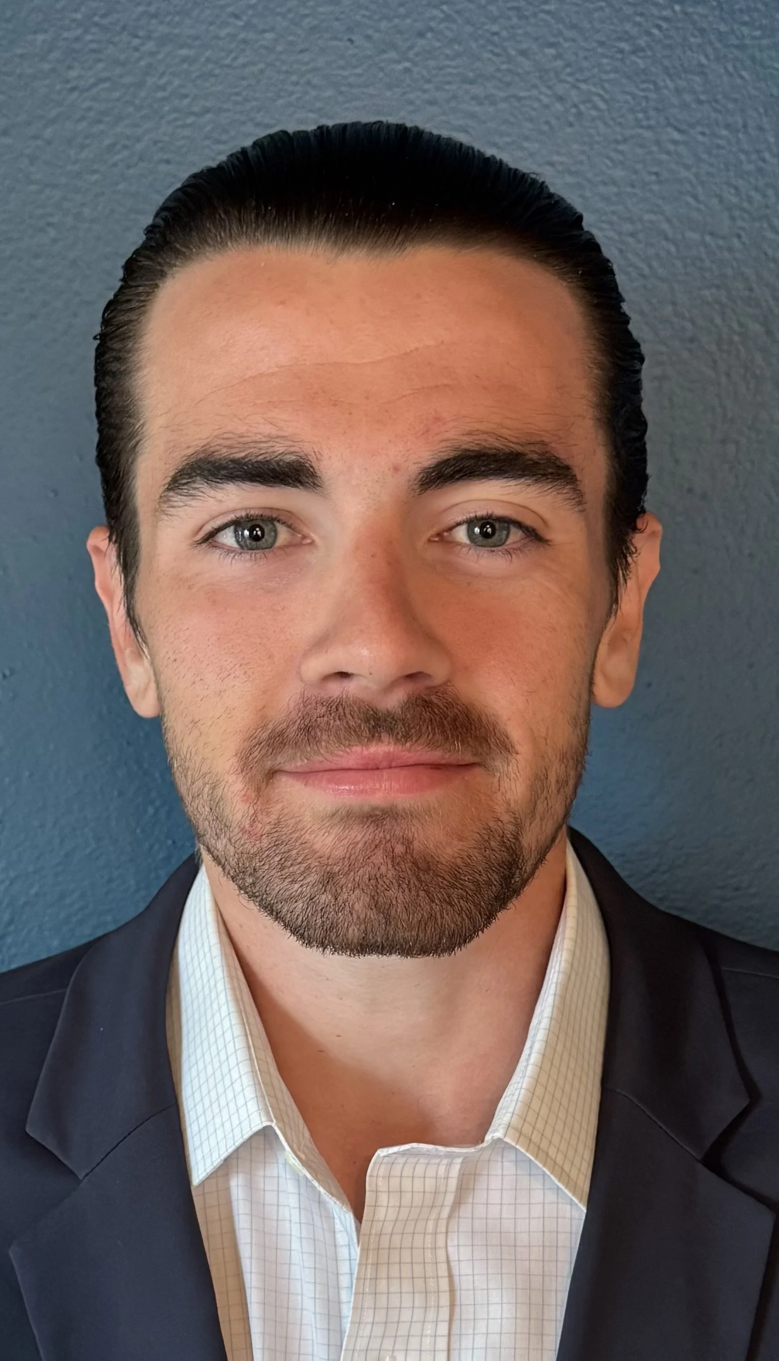 Portrait of a man with dark hair, blue eyes, and a beard, wearing a dark blazer and light checkered shirt, standing against a blue textured wall.