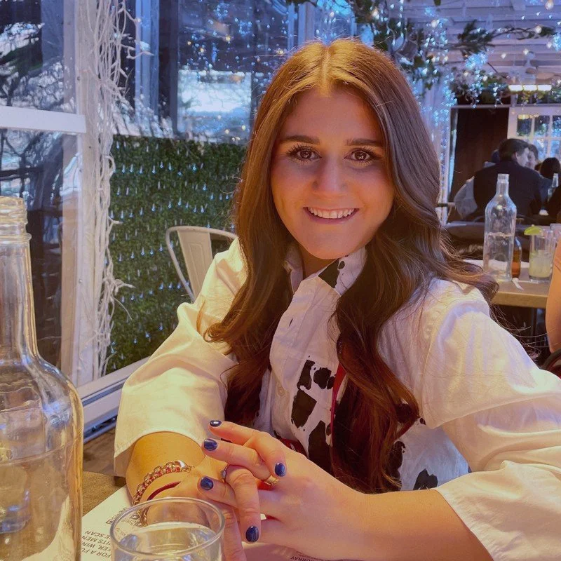 A young woman with long brown hair and a big smile, sitting at a restaurant table. She is wearing a white shirt with black patterns and has manicured nails painted dark blue. There are bottles and glasses on the table, and the background shows a decorated outdoor or indoor dining area with string lights and other people dining.