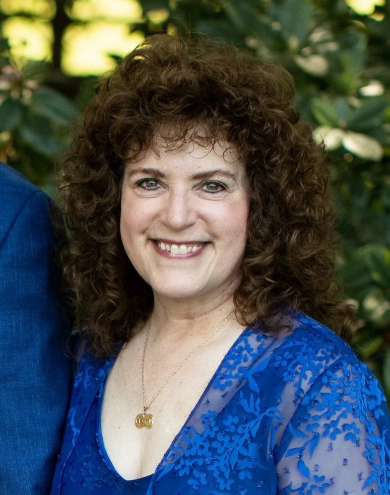 A woman with curly brown hair smiling, wearing a blue dress with lace details, and a gold necklace with a pendant, standing outdoors with green foliage in the background.