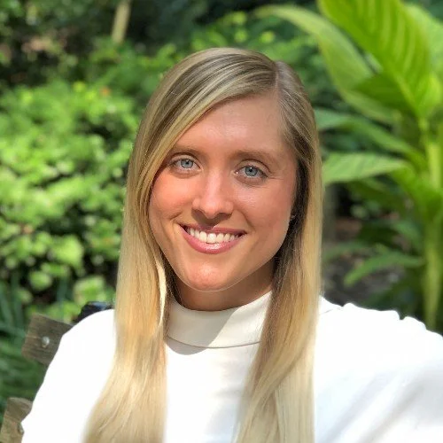 A young woman with long blonde hair and blue eyes smiling outdoors in front of green foliage.