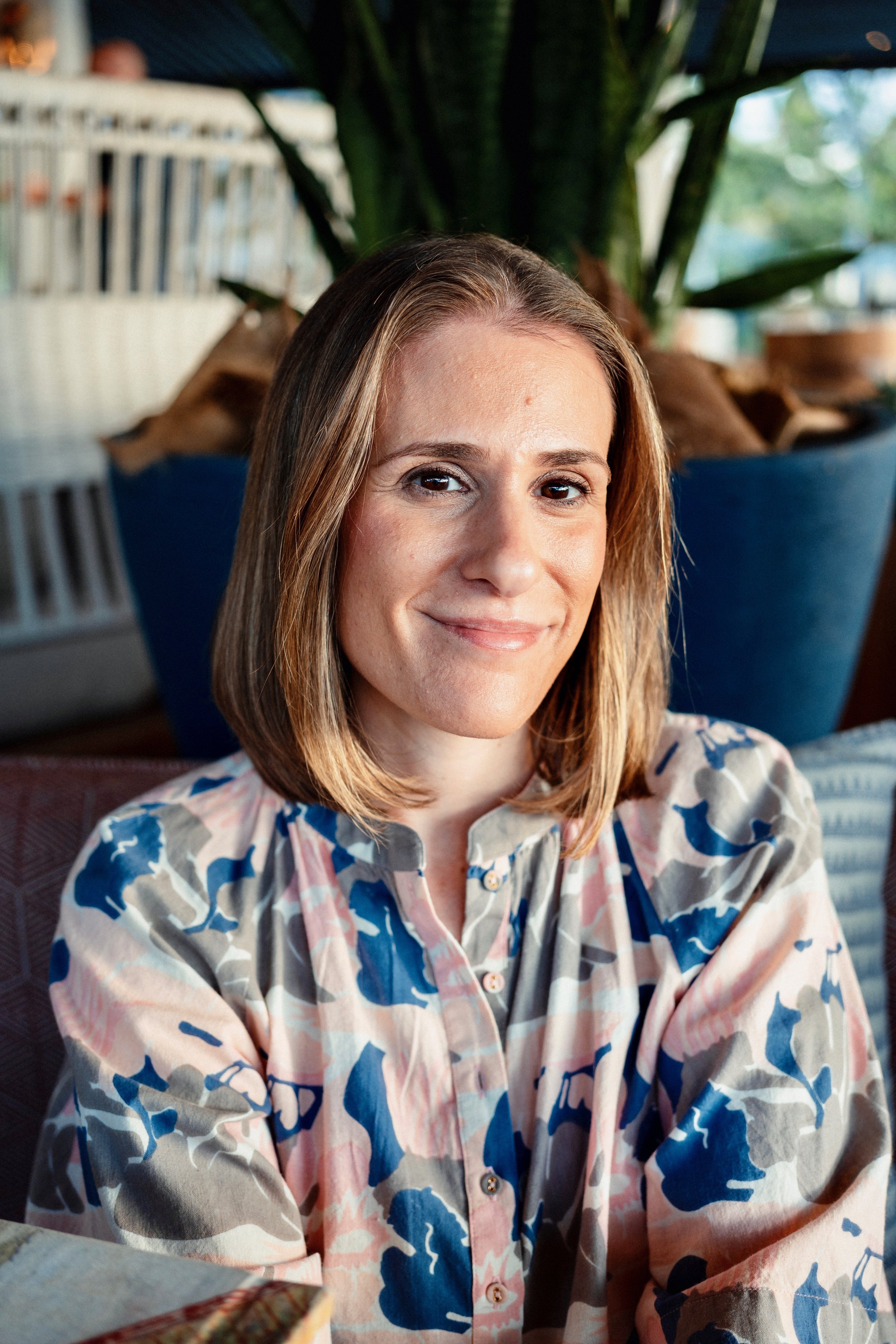 A woman with shoulder-length light brown hair smiling at the camera, wearing a colorful patterned blouse, sitting indoors near a green plant.