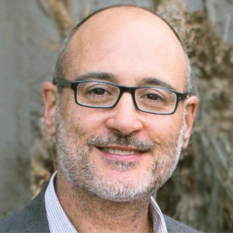 Close-up of a smiling middle-aged man with glasses, graying beard, and bald head, wearing a collared shirt and blazer, outdoors with blurred natural background.