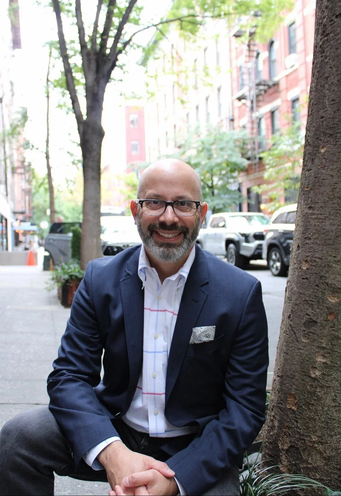 Man, outside on city street, wearing blue blazer and white striped dress shift.  He is wearing glasses, smiling.