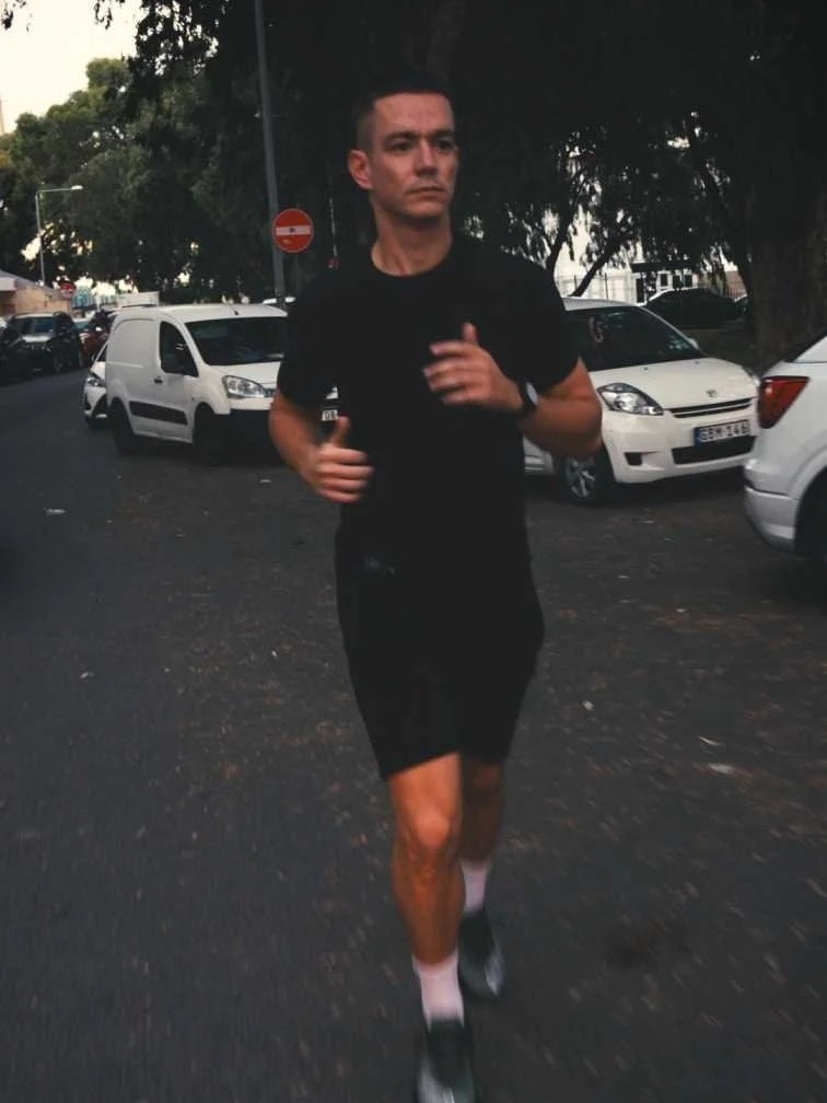A man jogging on a street at dusk, wearing black athletic clothing, with parked cars and trees in the background.