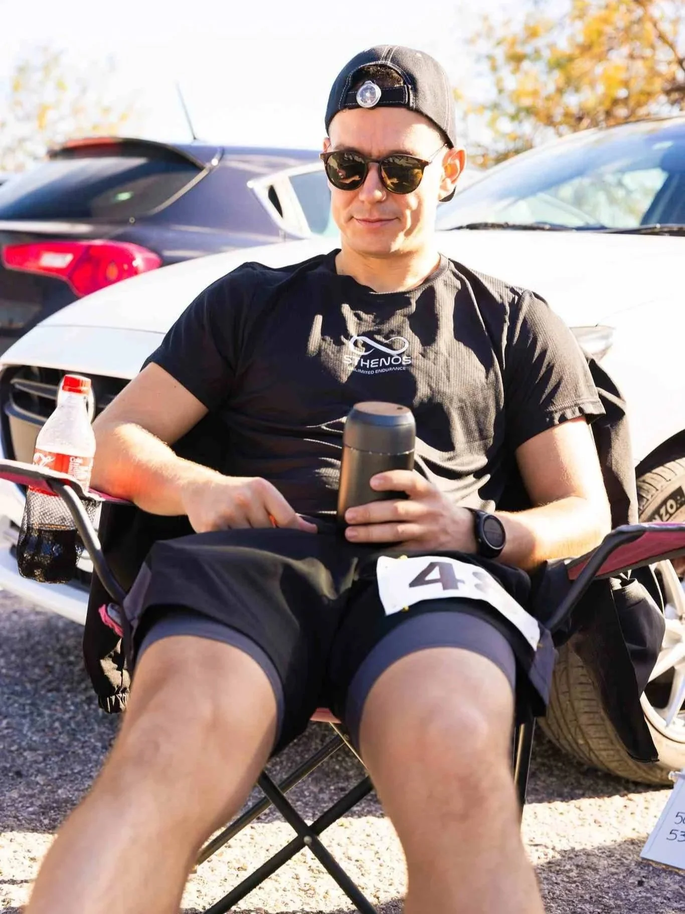 A man sitting outdoors in a folding chair, wearing sunglasses, a black cap, black shorts, and a black T-shirt, holding a travel mug, with cars parked behind him.