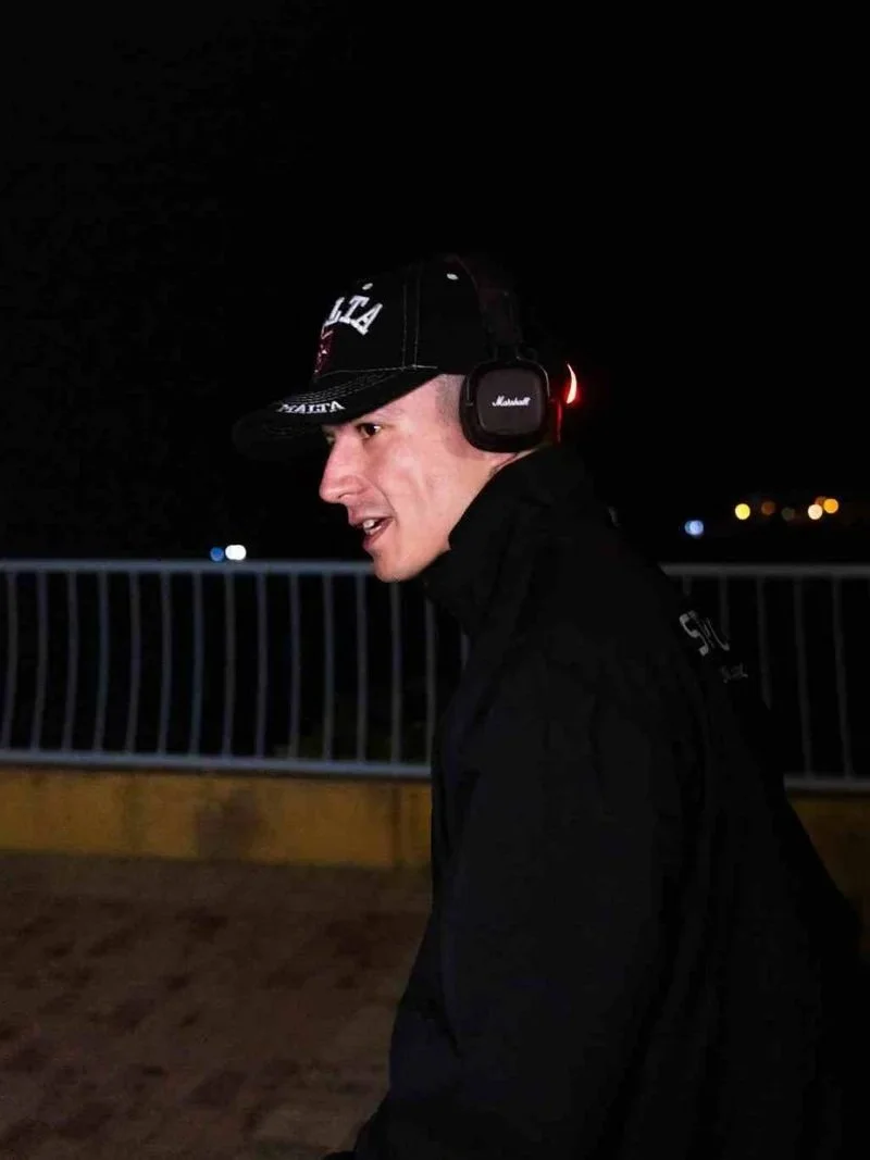 A young man wearing a black baseball cap, black jacket, and Marshall headphones is walking outdoors at night with a dark sky and city lights in the background.