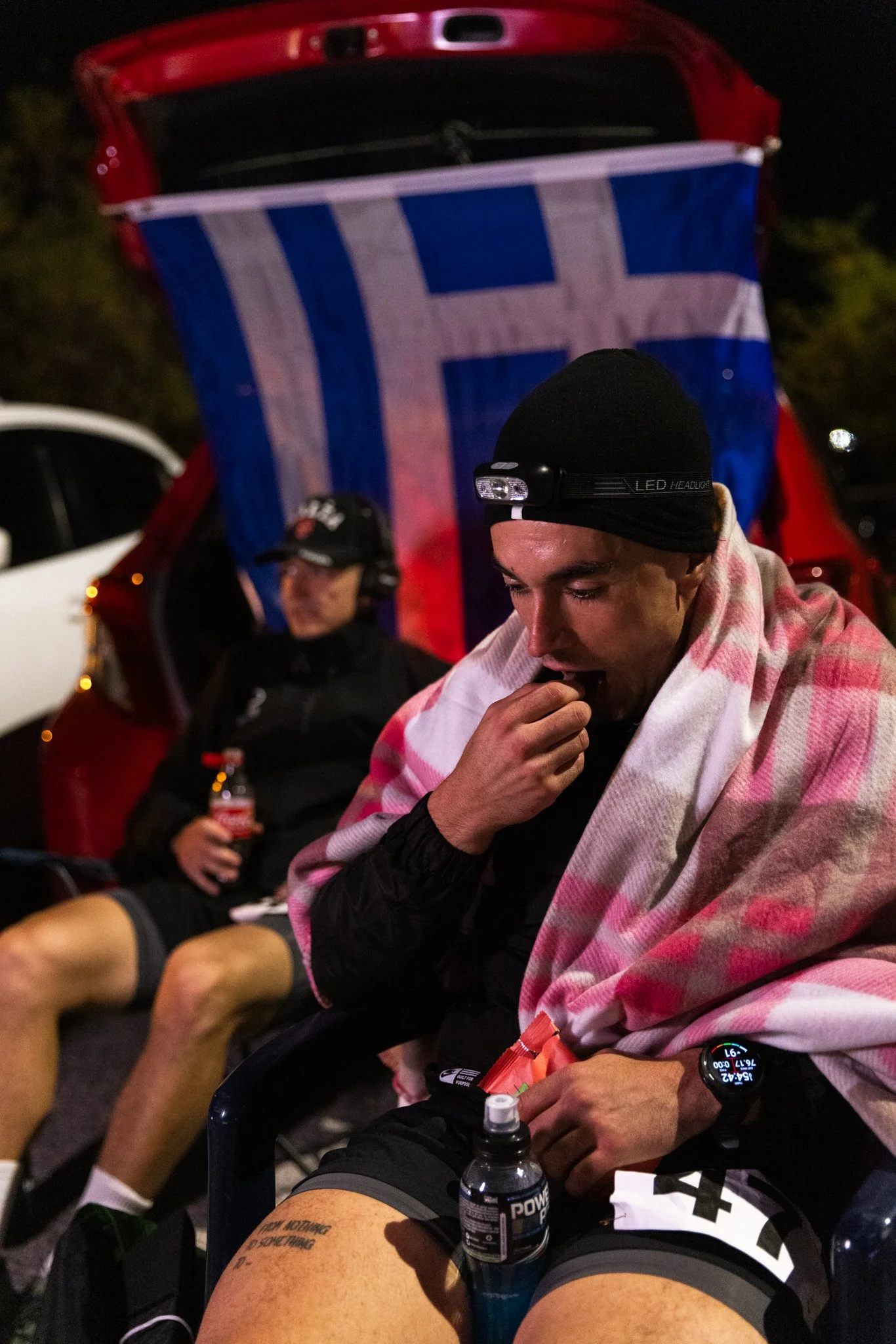 Two male athletes sitting outdoors at night, one wrapped in a pink and white blanket, wearing a black headlamp, with a tattoo on his thigh and a water bottle nearby, behind an open trunk with a blue and white flag hanging inside.