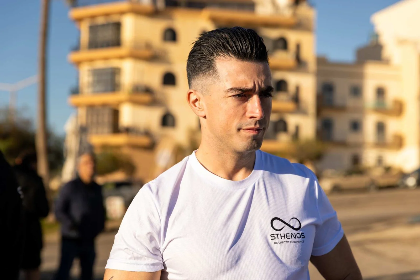 A man in a white T-shirt with the logo 'STHENOS Unlimited Endurance' stands outdoors with a modern building in the background, during daylight.