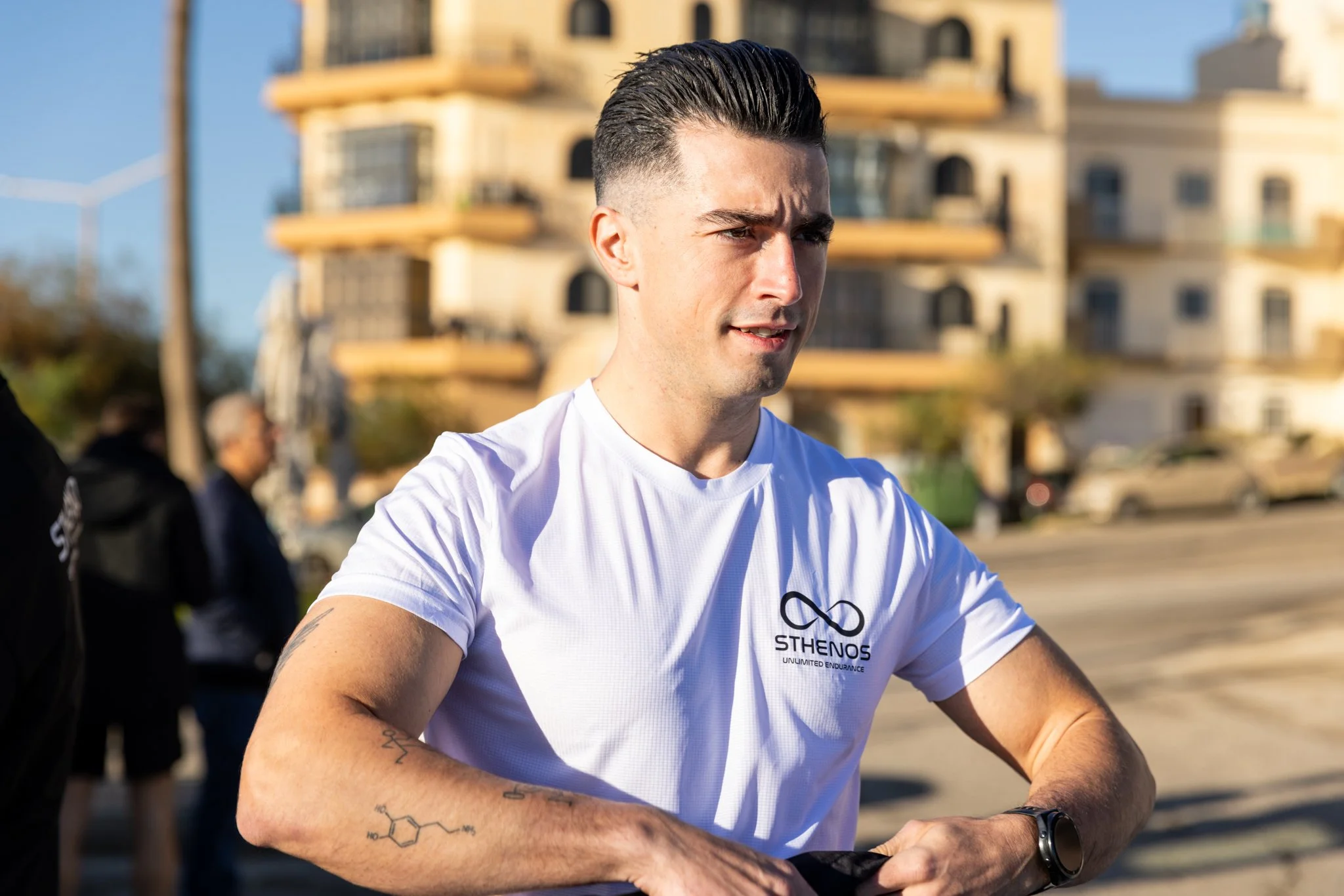 A young man in a white t-shirt with the logo 'Sthenos' and the phrase 'Unlimited Endurance' is outdoors in sunlight, absorbing sweat after a run or workout, with a modern building in the background.