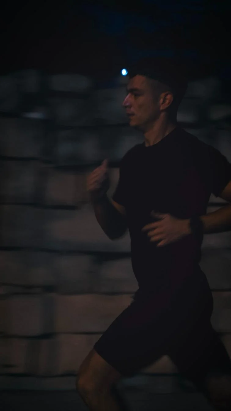 A man running outdoors at night, wearing a headlamp, black shirt, black shorts, and a wristwatch.