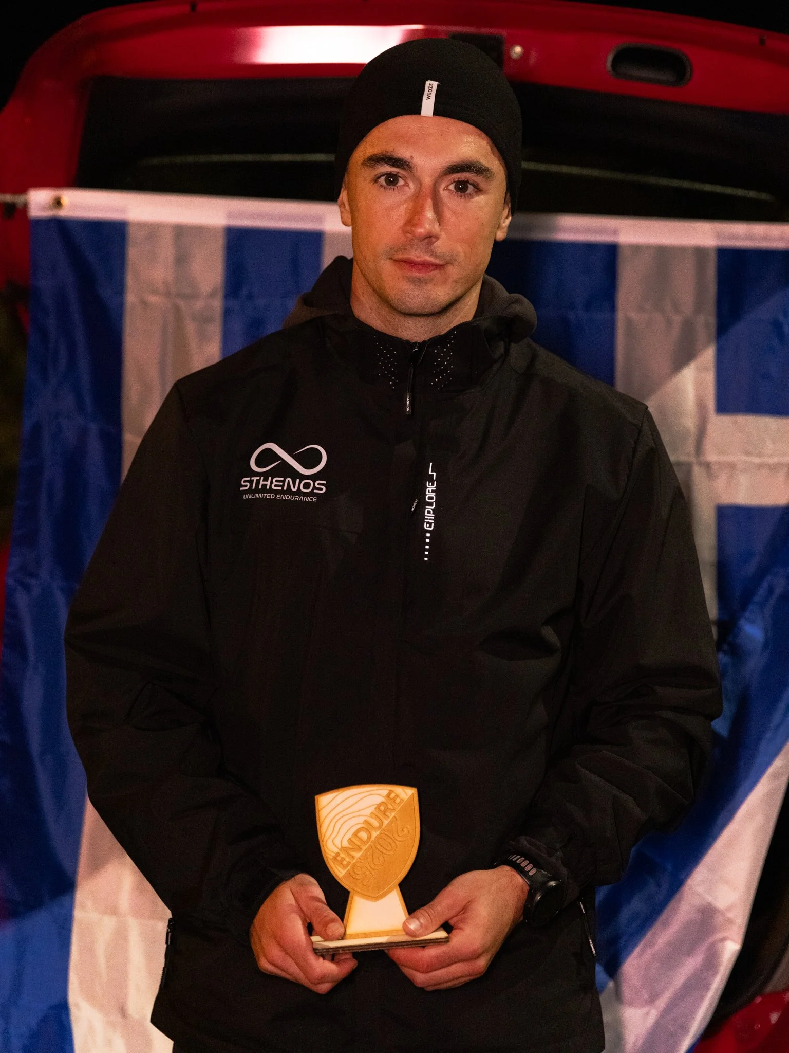A man dressed in black outdoor gear holding a wooden award with the word 'ENDURO' engraved on it. Behind him is a red vehicle and a flag with blue and white stripes.