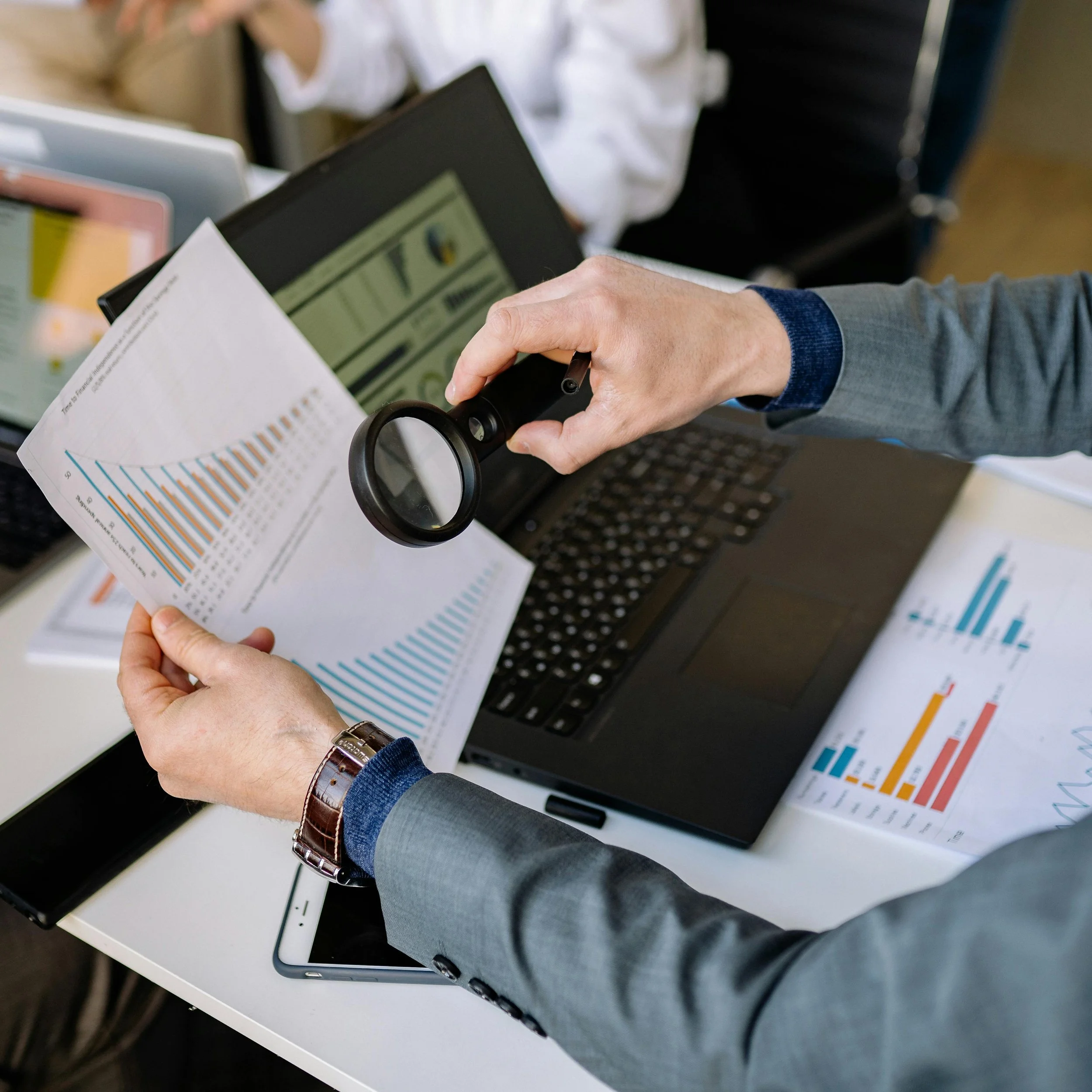 Person in a business suit examining financial reports with a magnifying glass, surrounded by a laptop and spreadsheets on a desk.