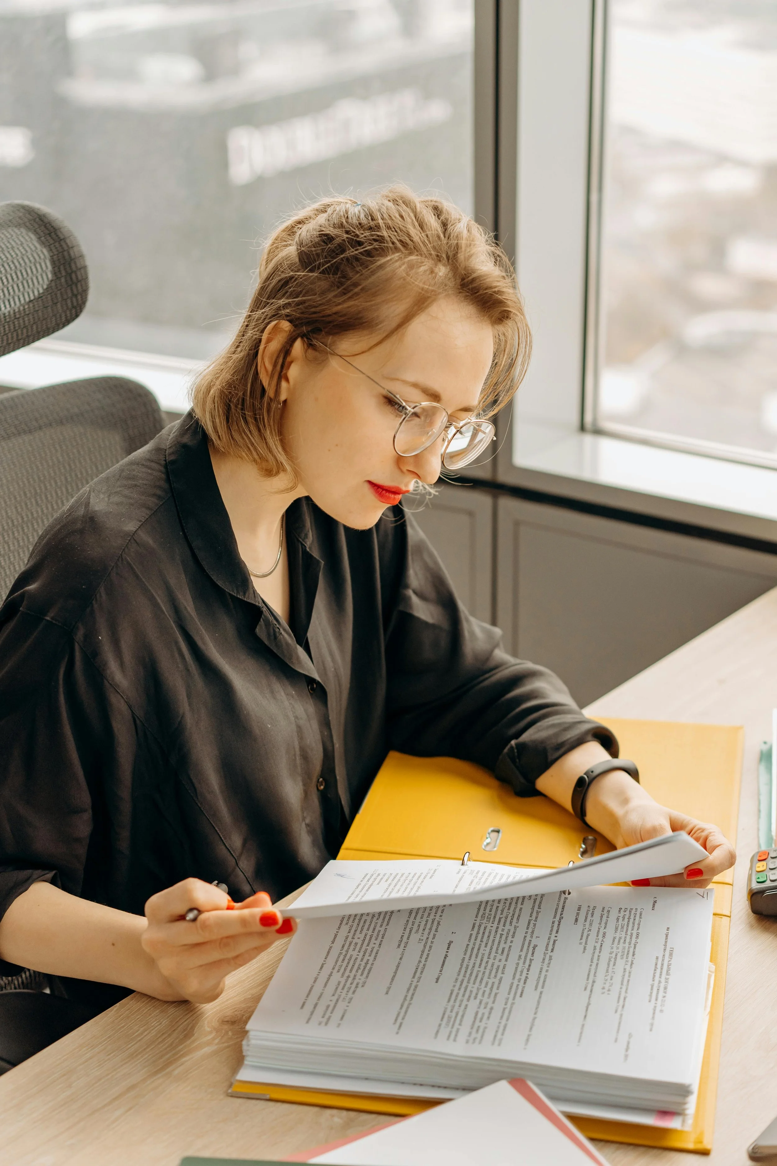 A woman with shoulder-length hair, wearing glasses and a black shirt, sitting at a desk with a large stack of papers, a yellow folder, and a calculator, in front of a window.