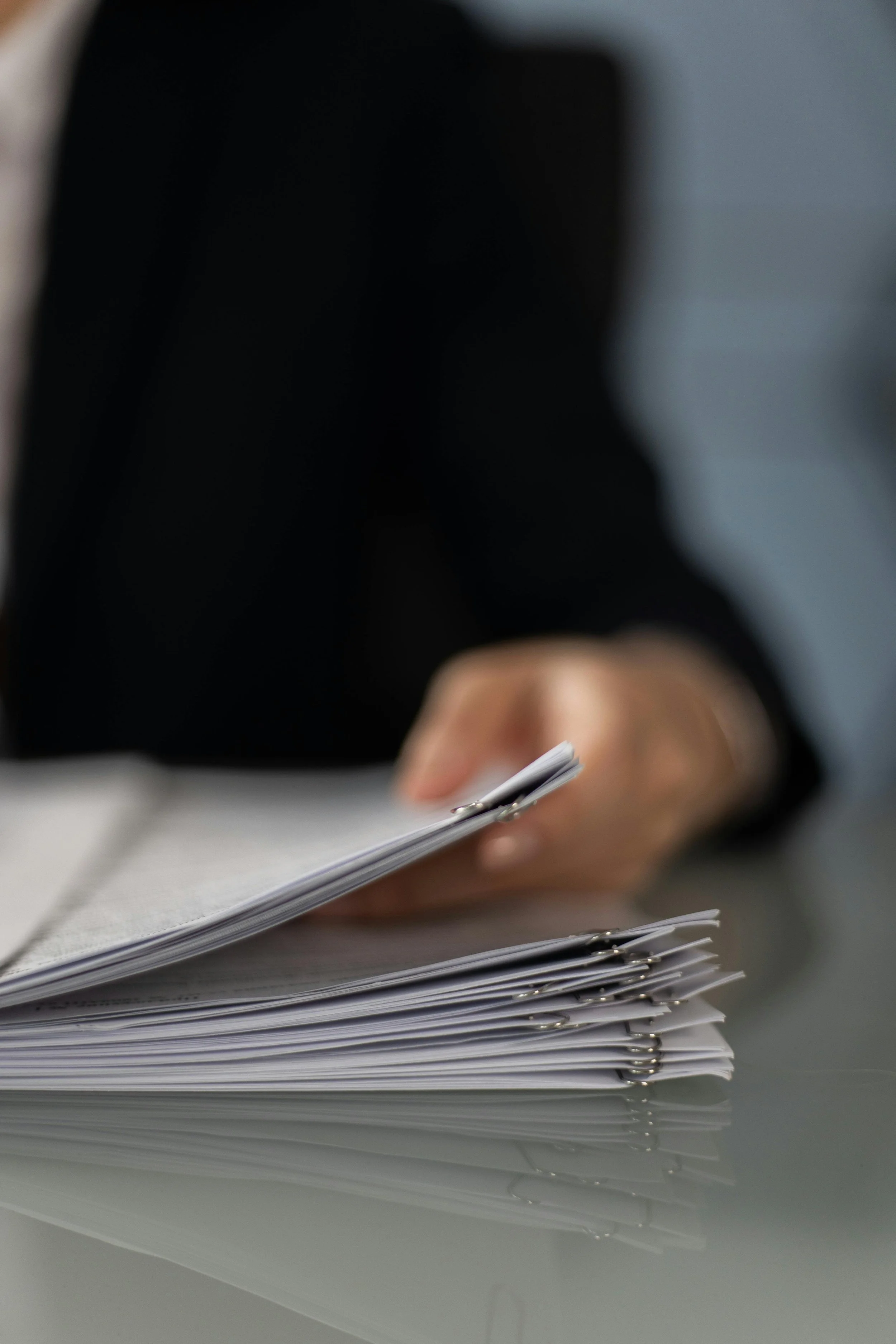A person in a black blazer is handling a stack of documents or papers, which are clipped together, on a reflective surface, with their hand in focus and the background blurred.