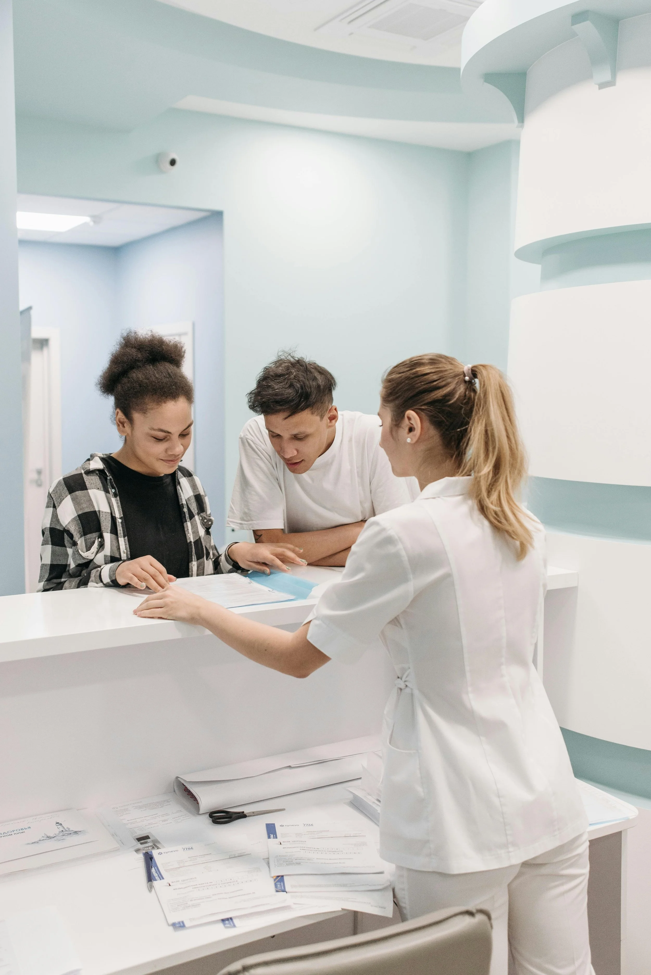 A nurse in white scrubs assisting two young patients at a reception desk in a medical office or clinic.