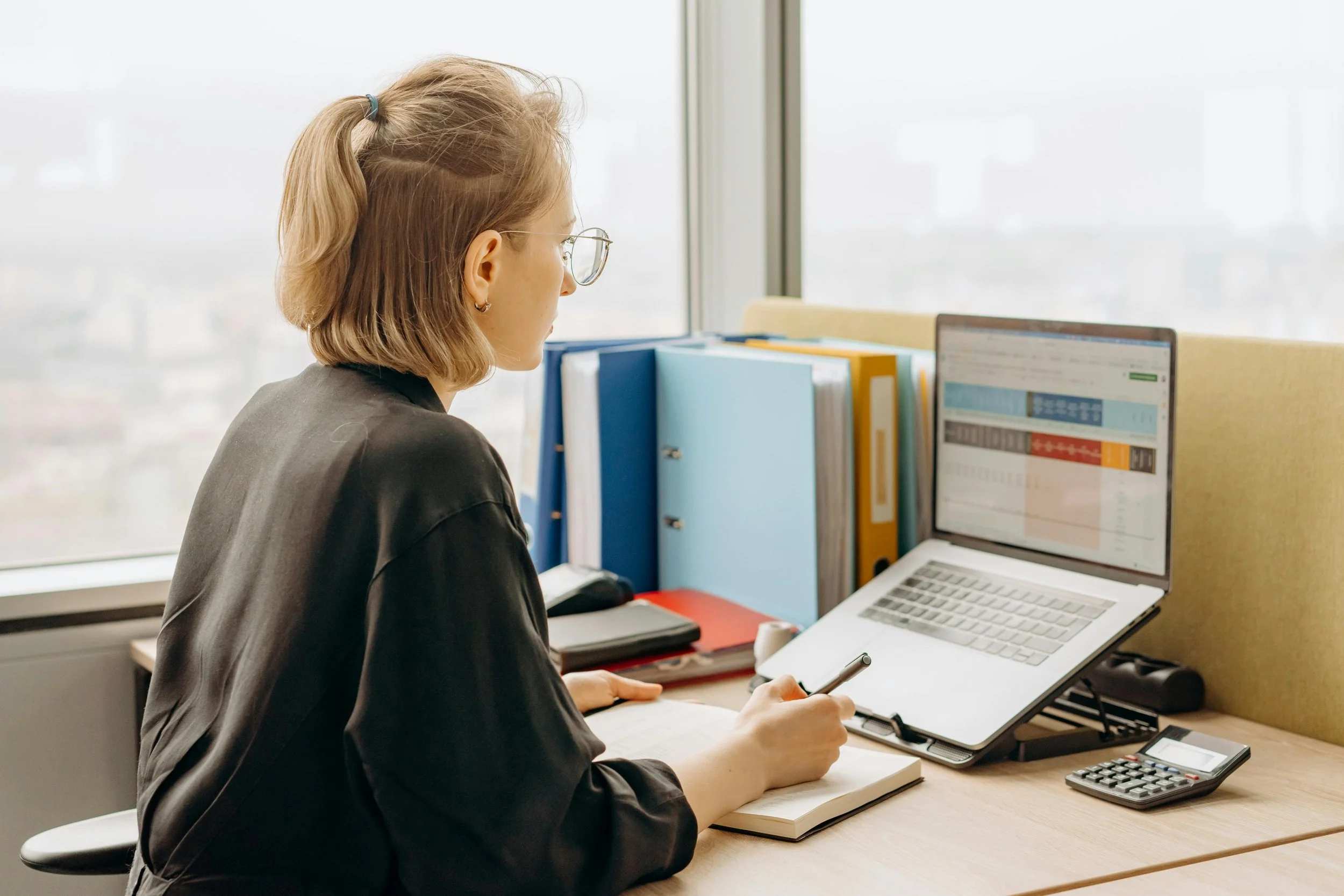 A woman with blond hair tied in a ponytail and wearing glasses, working at her desk with a laptop, notebooks, and file folders in an office.