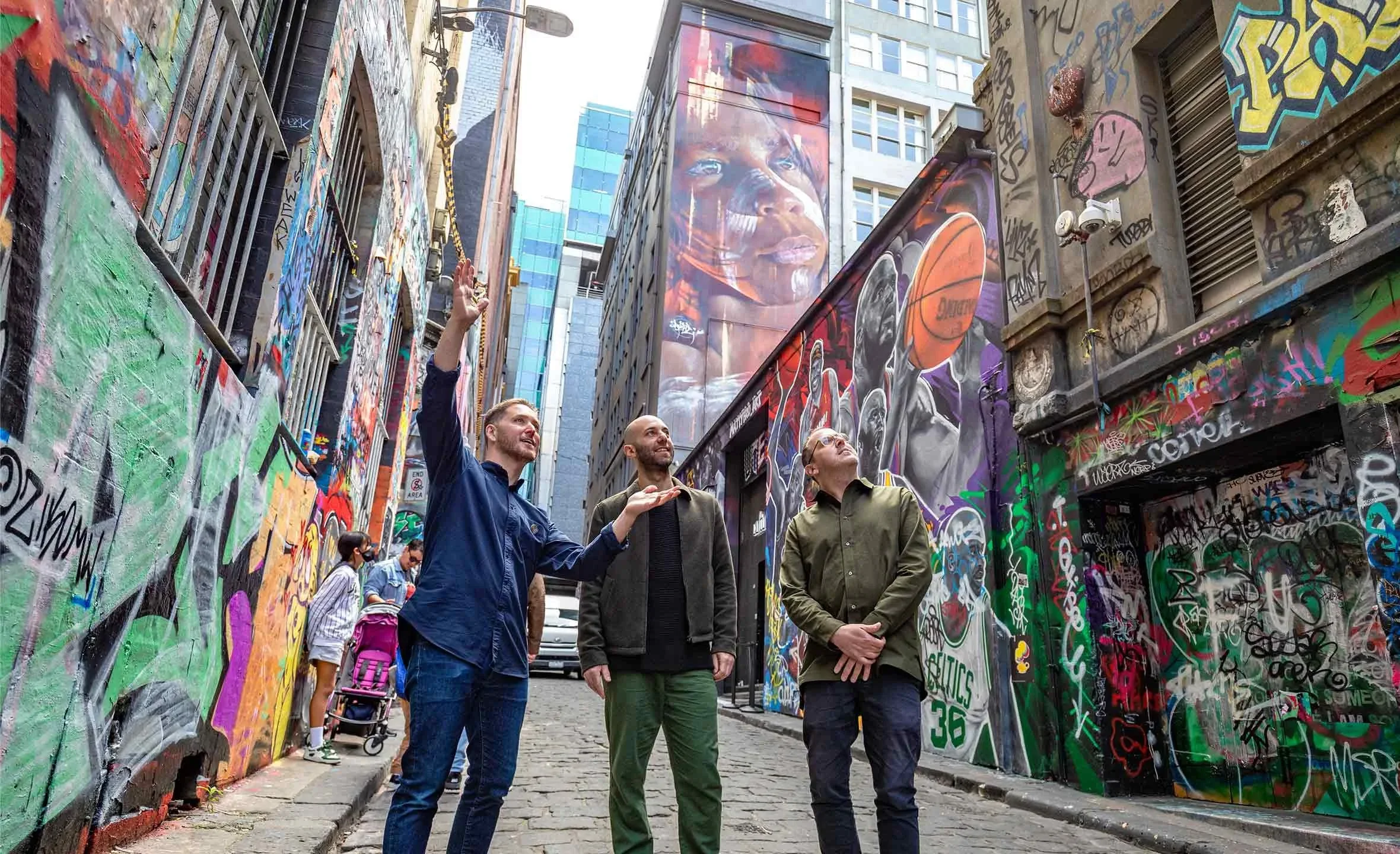 Three men are standing on a cobblestone street in a city alley with colorful graffiti and murals on the walls. One man is gesturing upward, while the others look on. There are a few pedestrians and a person with a pink backpack in the background.