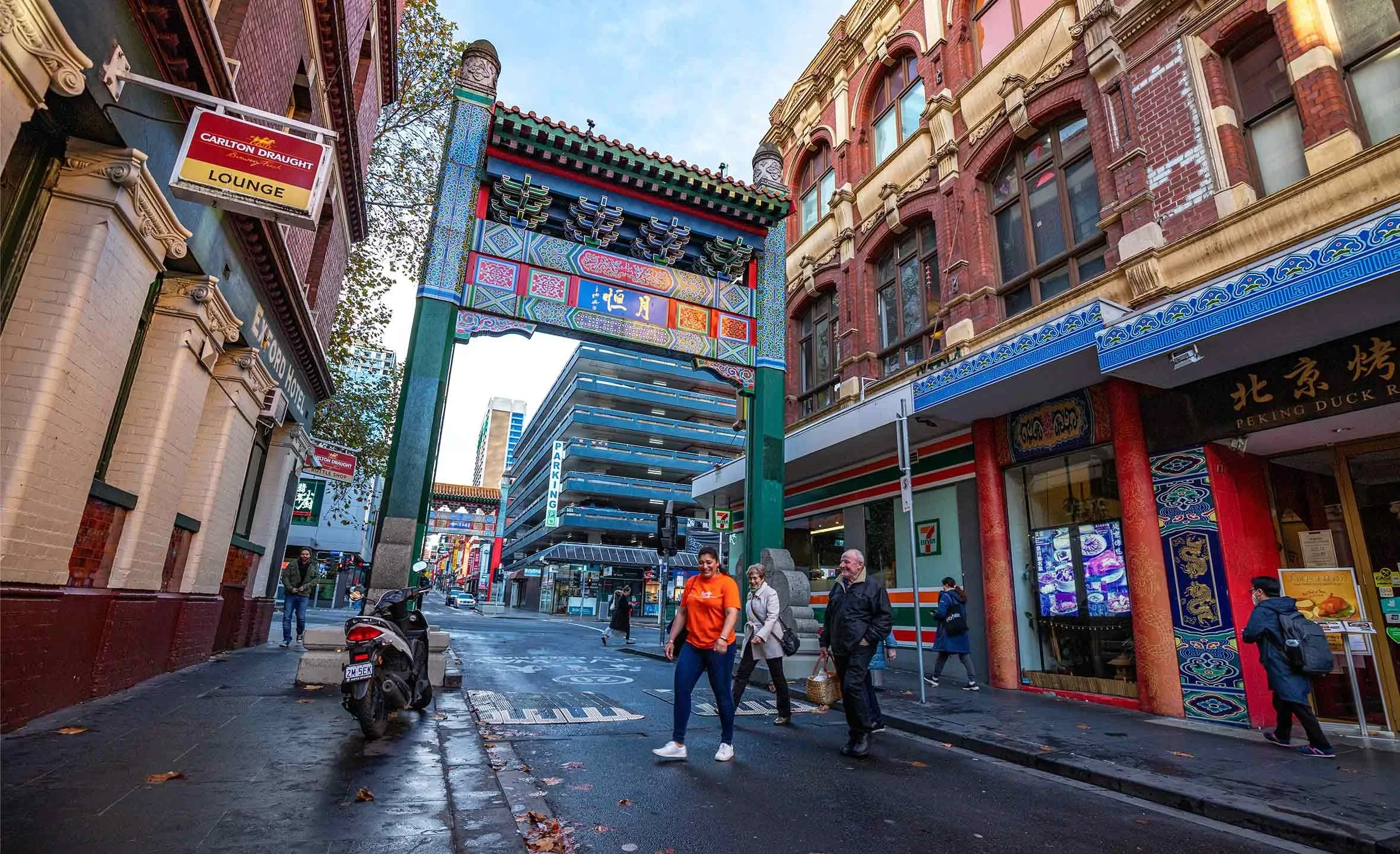 City street scene with a traditional Chinese archway, pedestrians, a motorcycle, and shops, including a 7-Eleven store.