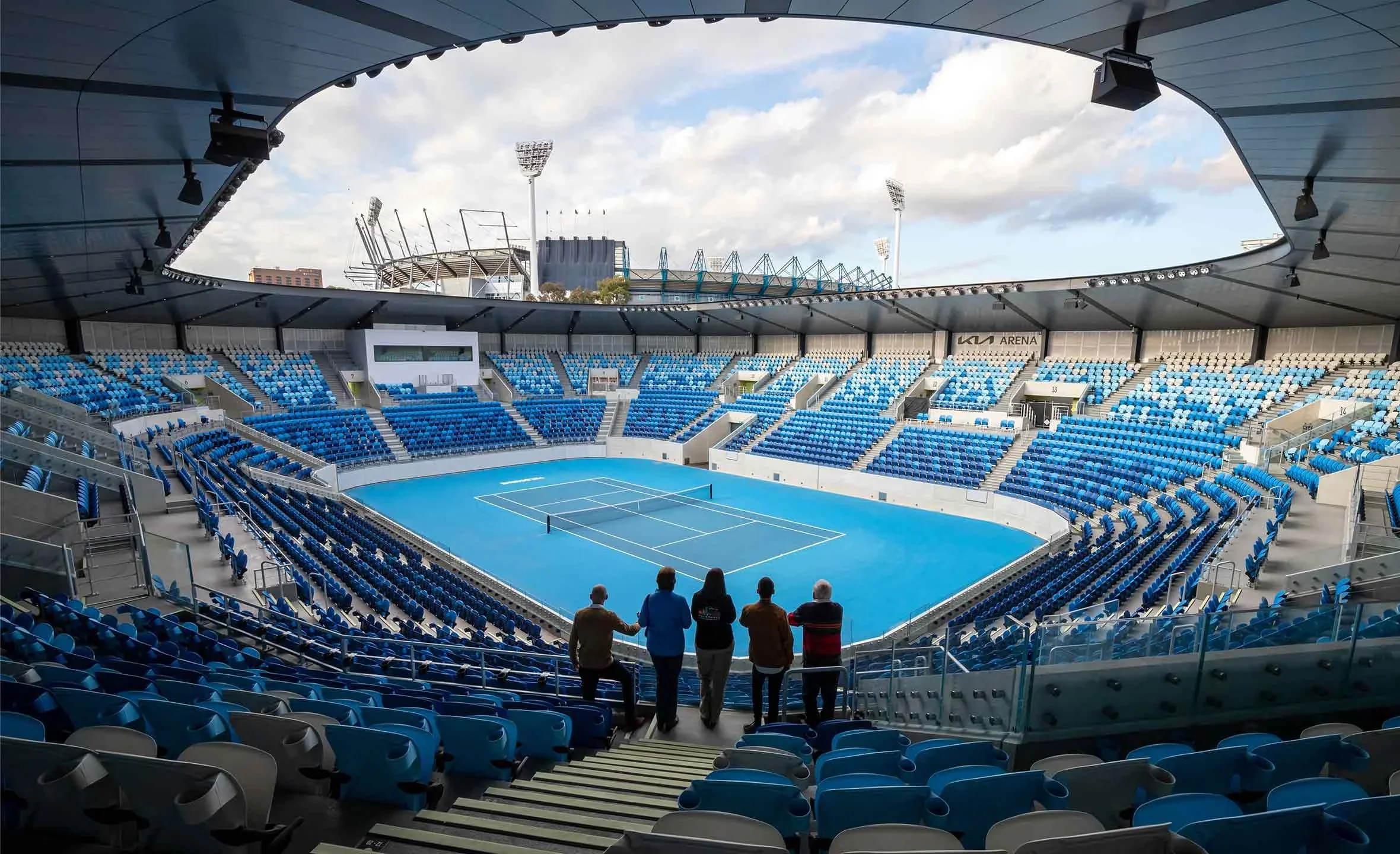 Empty blue tennis court inside a modern stadium with a partial view of people standing at the top rows, looking down at the court, under a partially open roof.