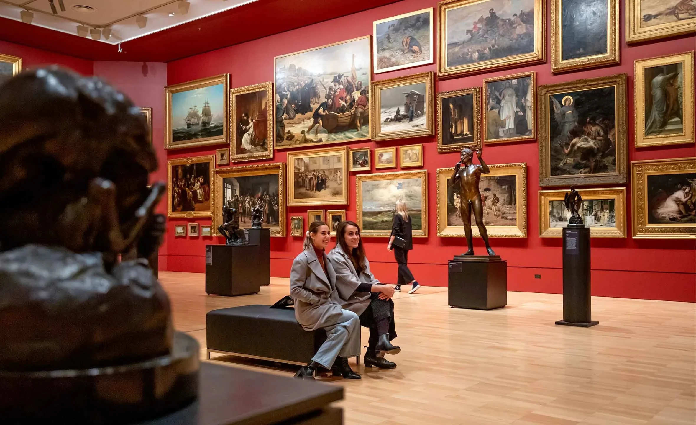 Two women sitting on a bench in an art gallery with red walls, surrounded by paintings and sculptures.