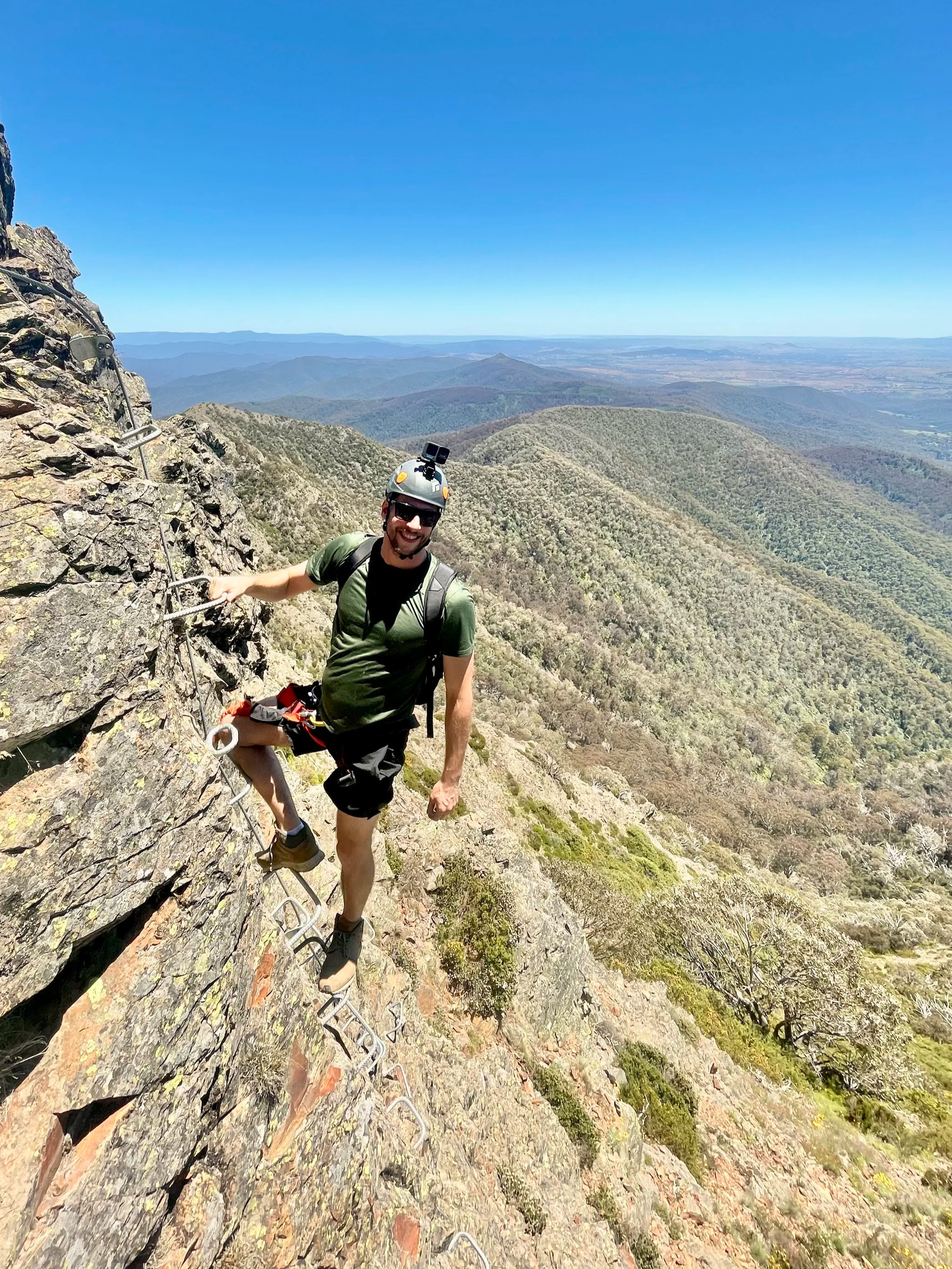 Justin Meneguzzi wearing a helmet and sunglasses while climbing a steep rocky mountain with metal rungs and safety cables, with a scenic view of green mountain ranges and a clear blue sky in the background.