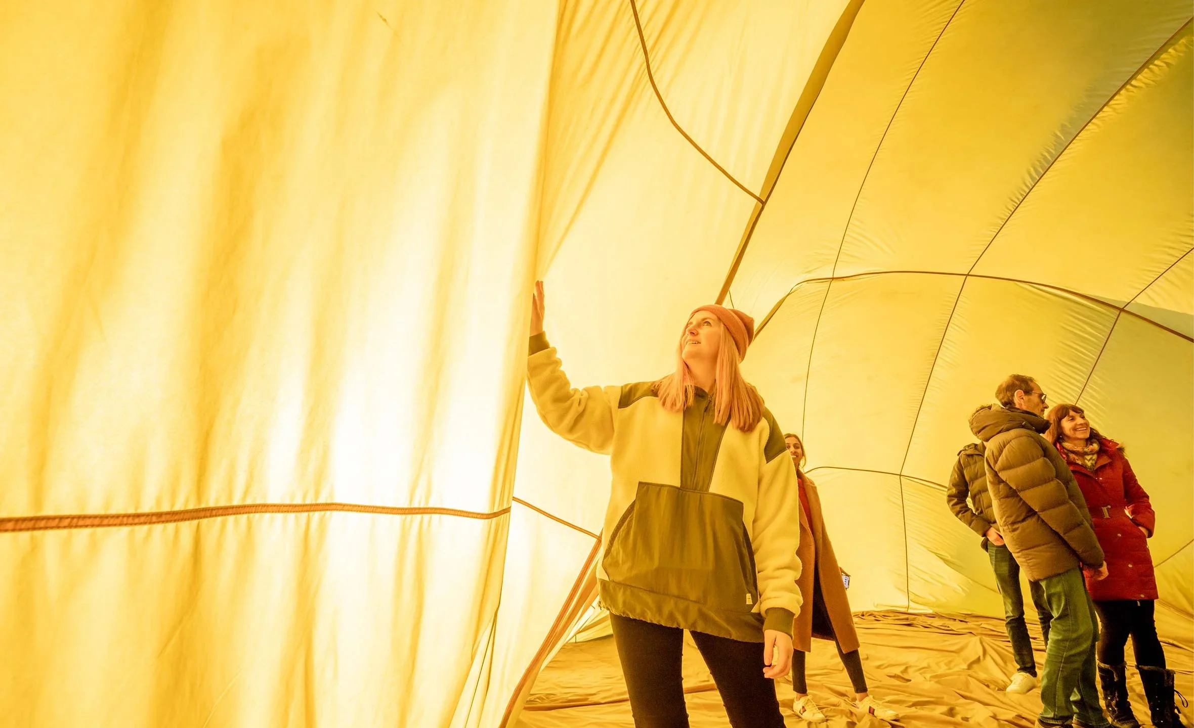 Group of people inside a large yellow hot air balloon envelope, one woman is looking up and touching the fabric, others are standing and talking.