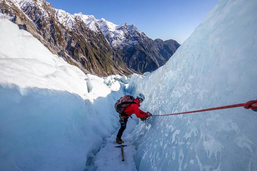 A person ice climbing on a narrow glacier crevasse with snow-covered mountains in the background.