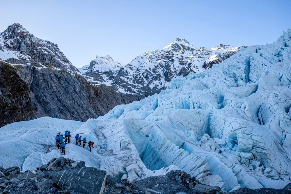 A group of climbers hiking on a glacier in a mountainous icy landscape with snow-covered peaks in the background.