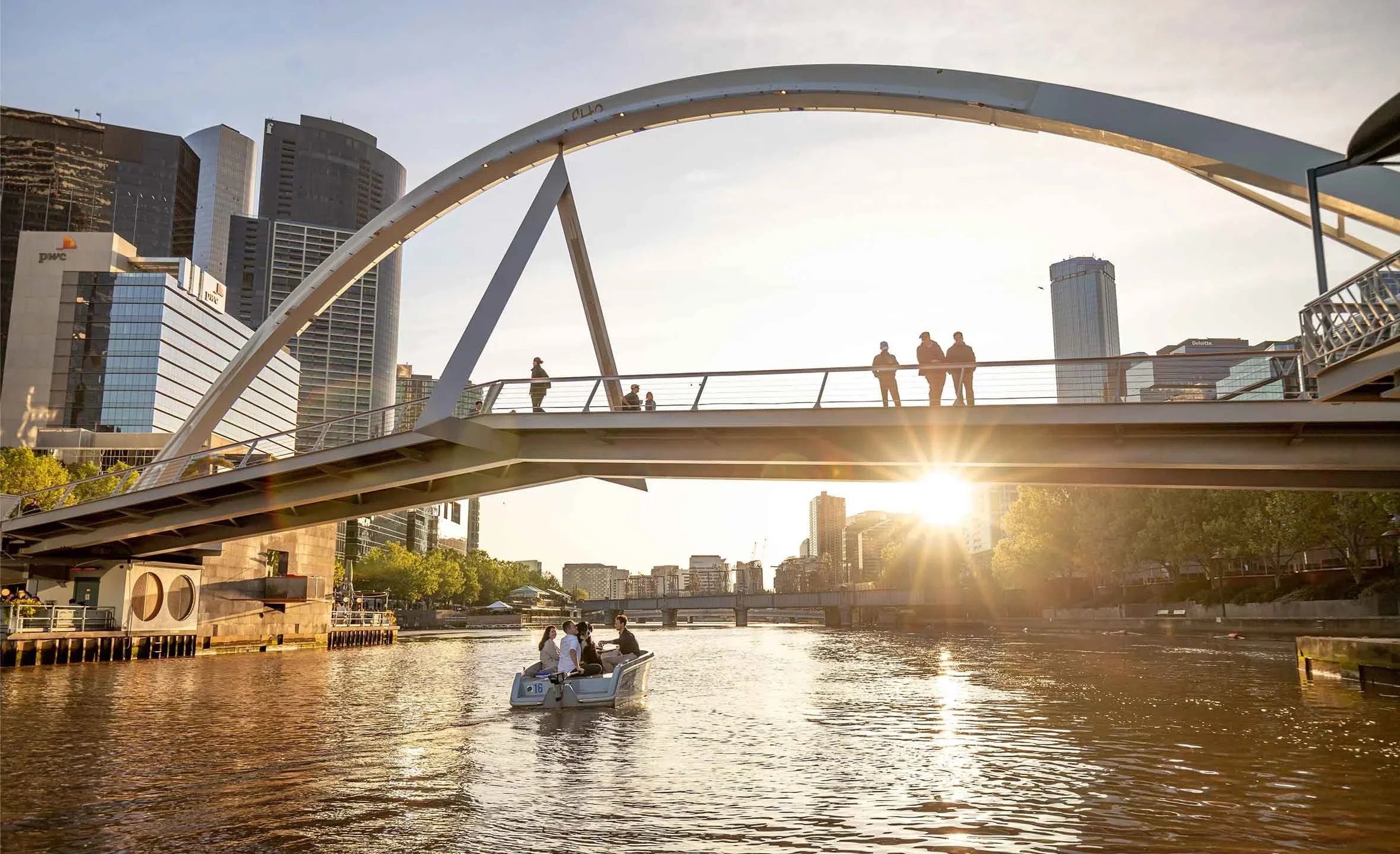 A modern cityscape with a river, a pedestrian bridge, and tall buildings in the background during sunset. People are walking on the bridge and two are riding a pedal boat on the water.