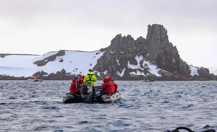 A small inflatable boat with four people wearing red and yellow jackets, sailing in a cold, snowy landscape with rocky cliffs and snow-covered land in the background.