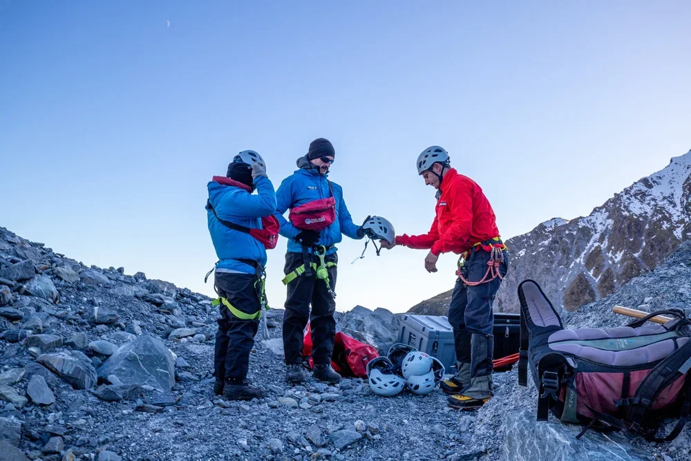Three hikers in gear on rocky mountain terrain, preparing helmets in a mountainous landscape with snow-capped peaks, during daylight.