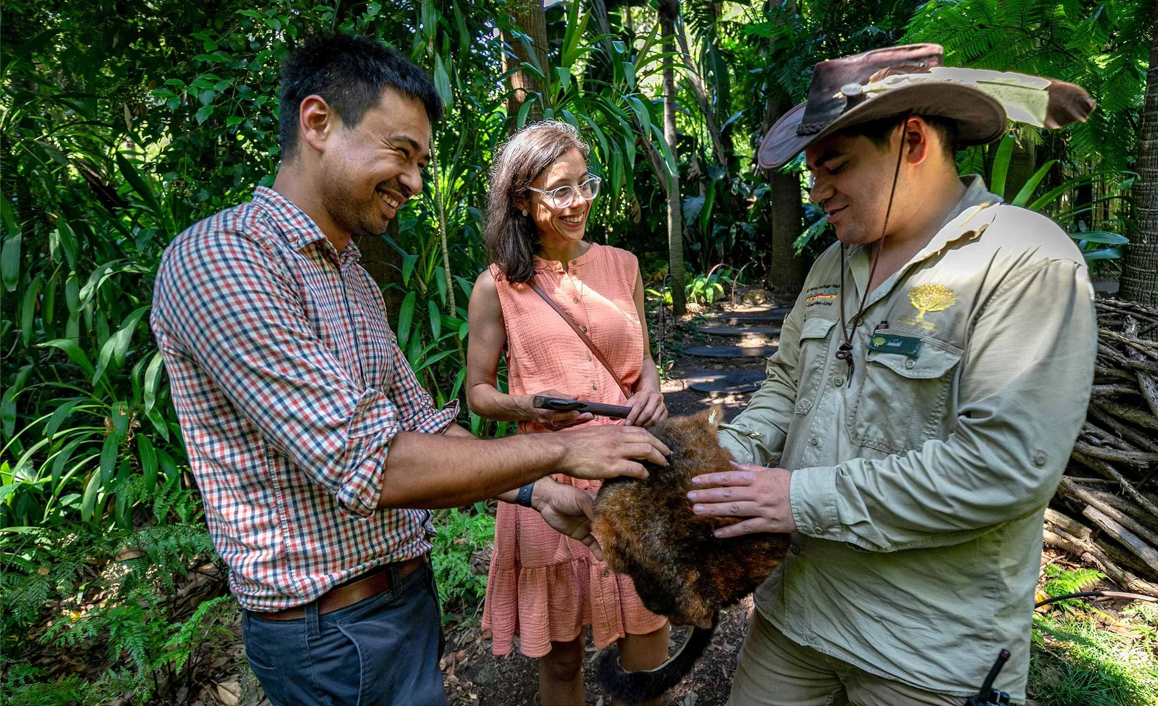 Three people in a lush forest with dense green foliage, one man in a checked shirt and one woman in a sleeveless dress observing a man in a ranger uniform holding a small animal in his hands.