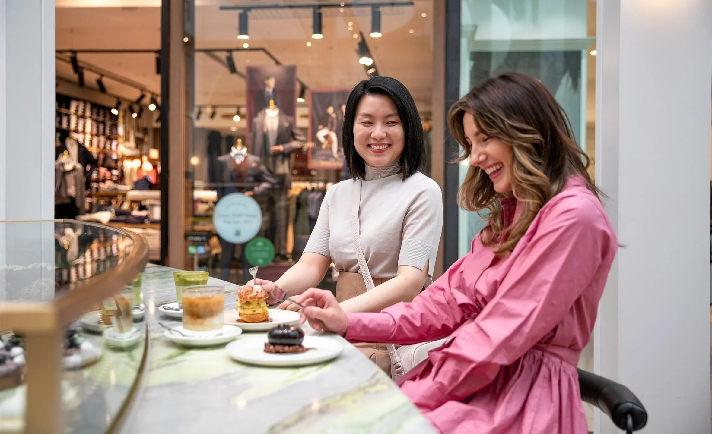 Two women sitting at a counter in a bakery or cafe, smiling and enjoying desserts, with a clothing store in the background.
