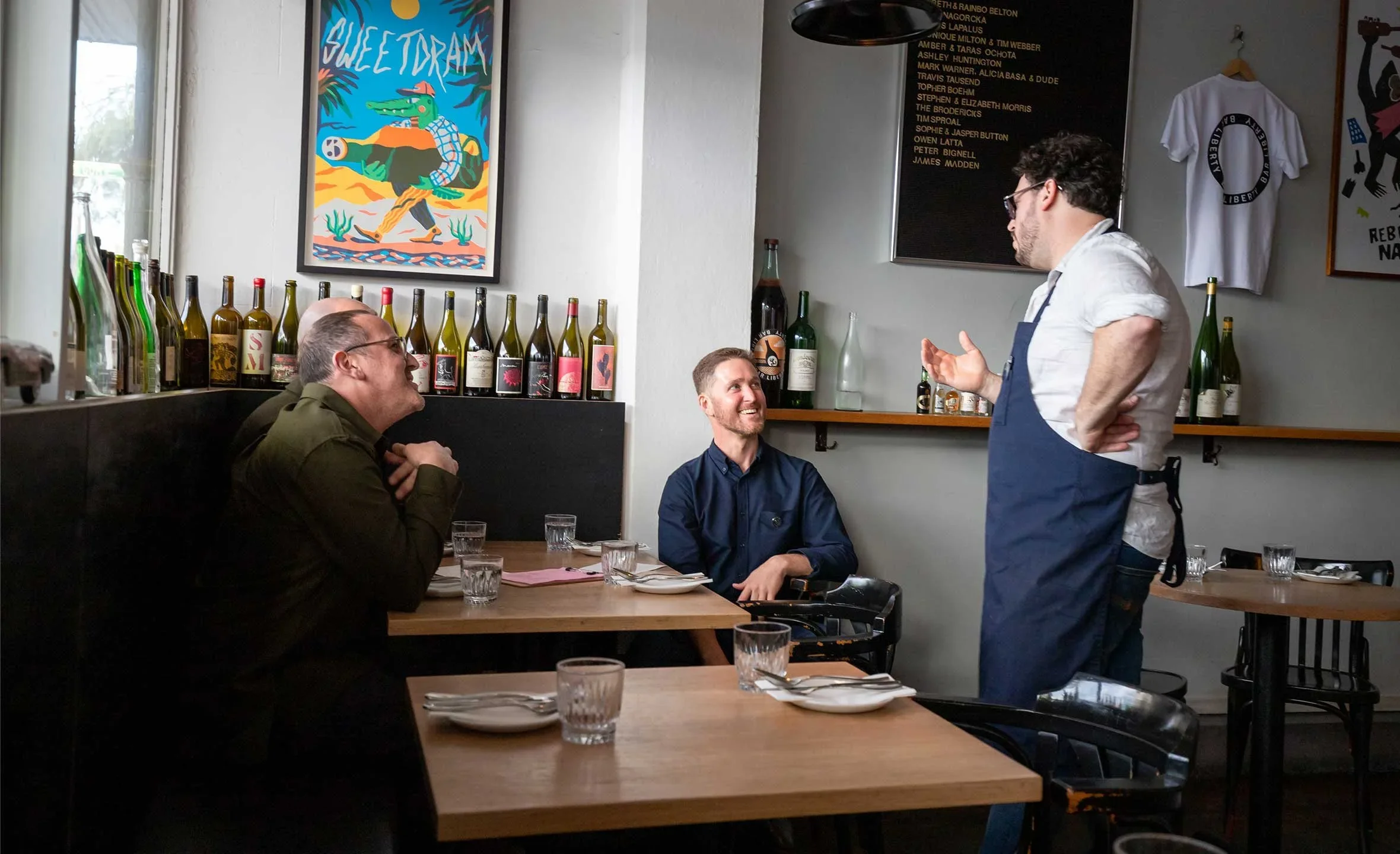 A waiter in a white shirt and blue apron talking to two male customers in a restaurant, with one wearing glasses and the other looking up and smiling. The restaurant has bottles and artwork on the walls.